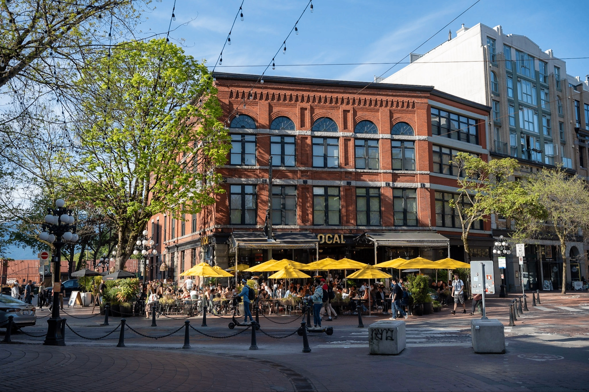 Outdoor café with yellow umbrellas and people sitting and walking near a red brick building on a sunny day.