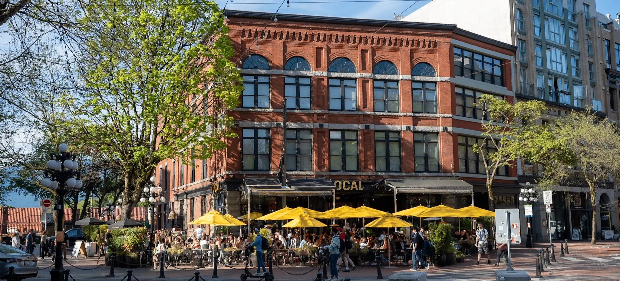 Outdoor café with yellow umbrellas and people sitting and walking near a red brick building on a sunny day.