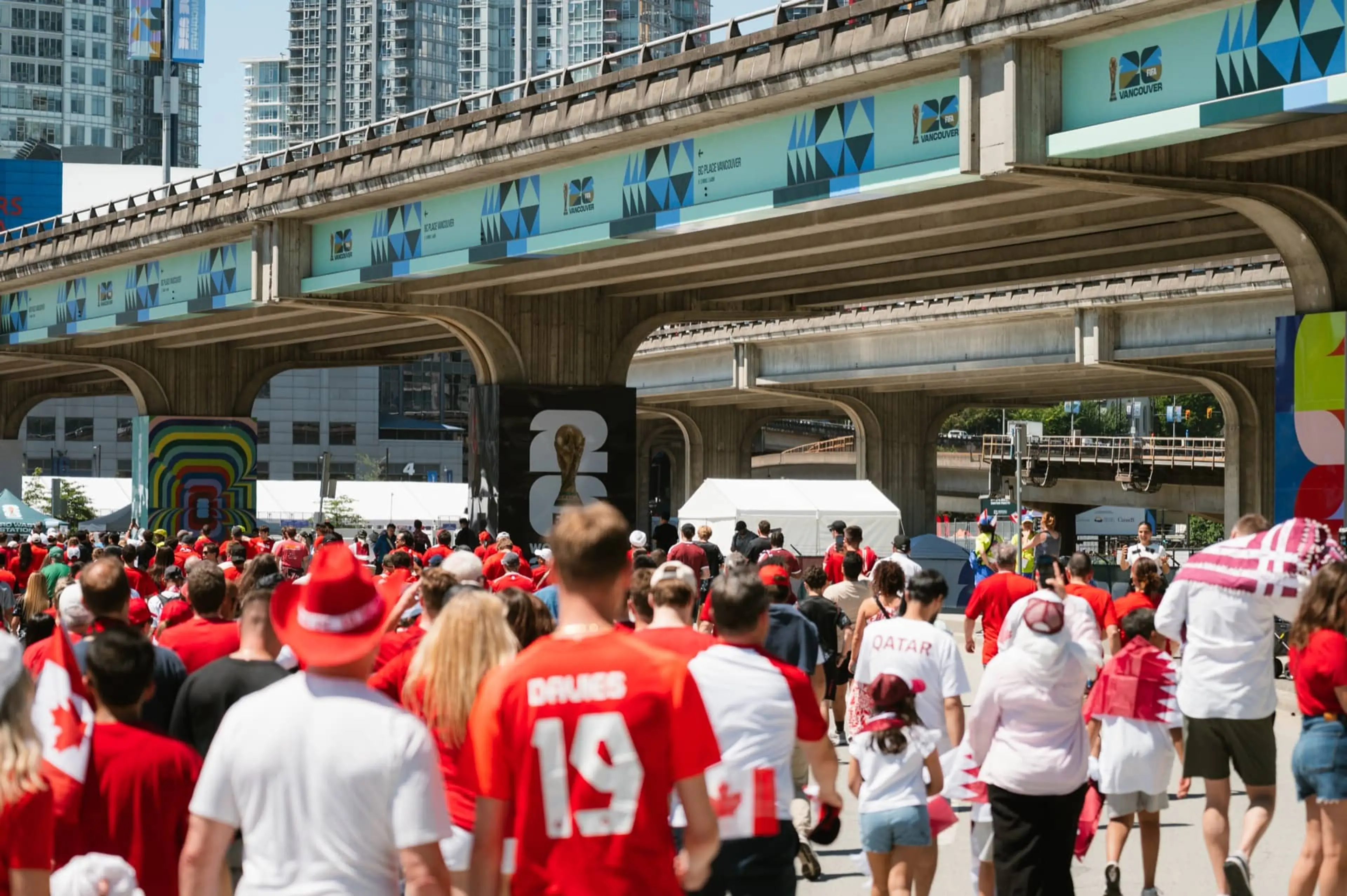 Crowd of people wearing red and white, some with Canadian flags, walking under a concrete overpass in a city.