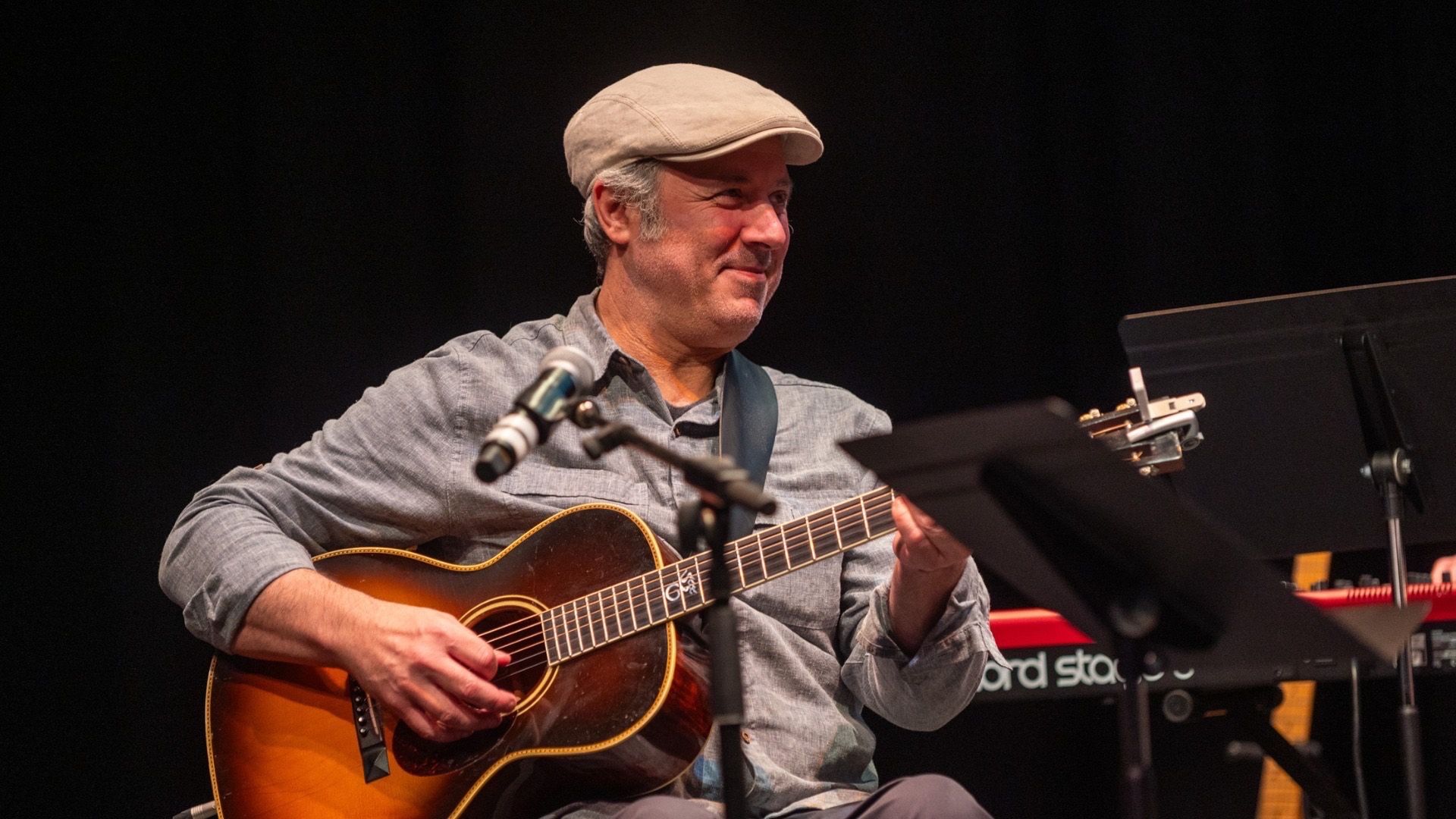 Person wearing a light gray shirt and beige cap playing an acoustic guitar on stage with a microphone and music stand nearby.