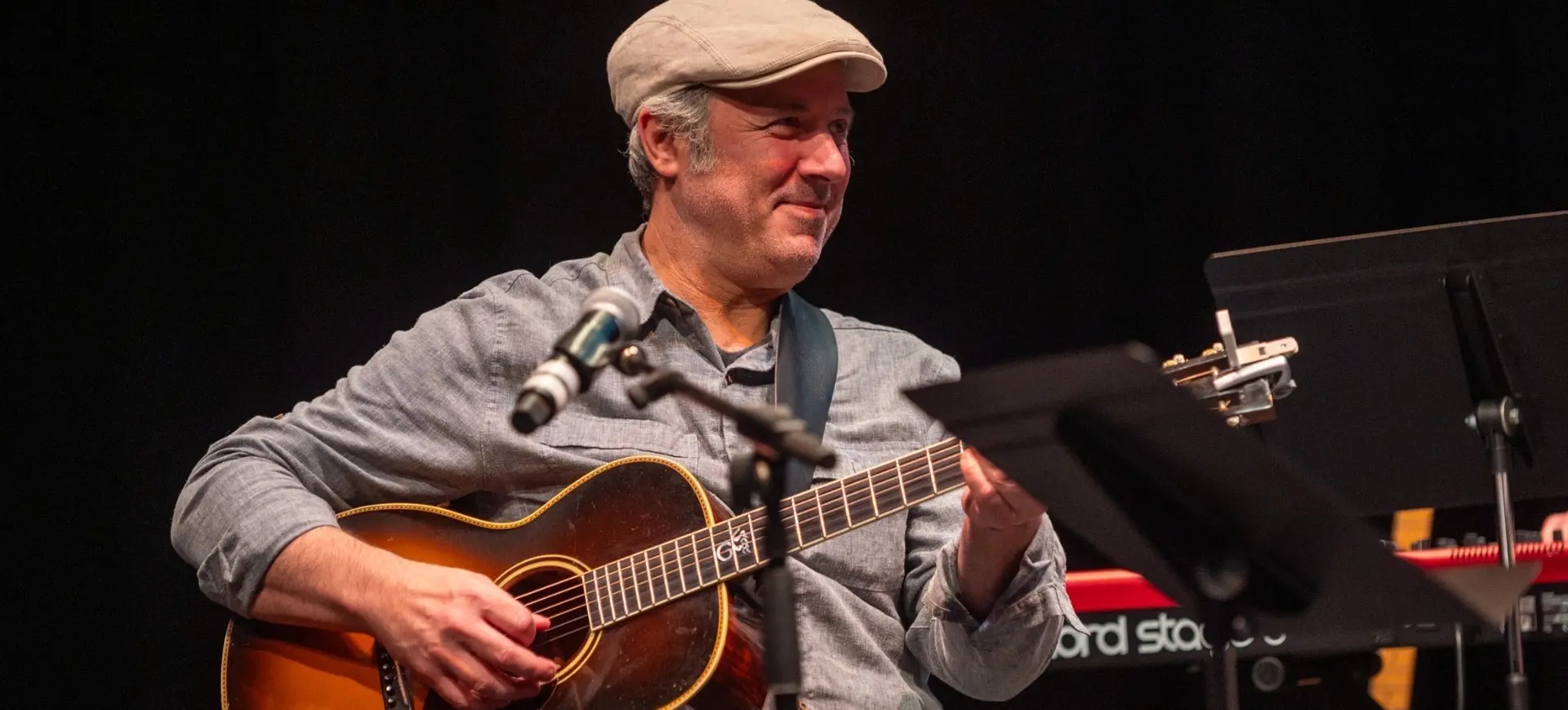 Person wearing a light gray shirt and beige cap playing an acoustic guitar on stage with a microphone and music stand nearby.