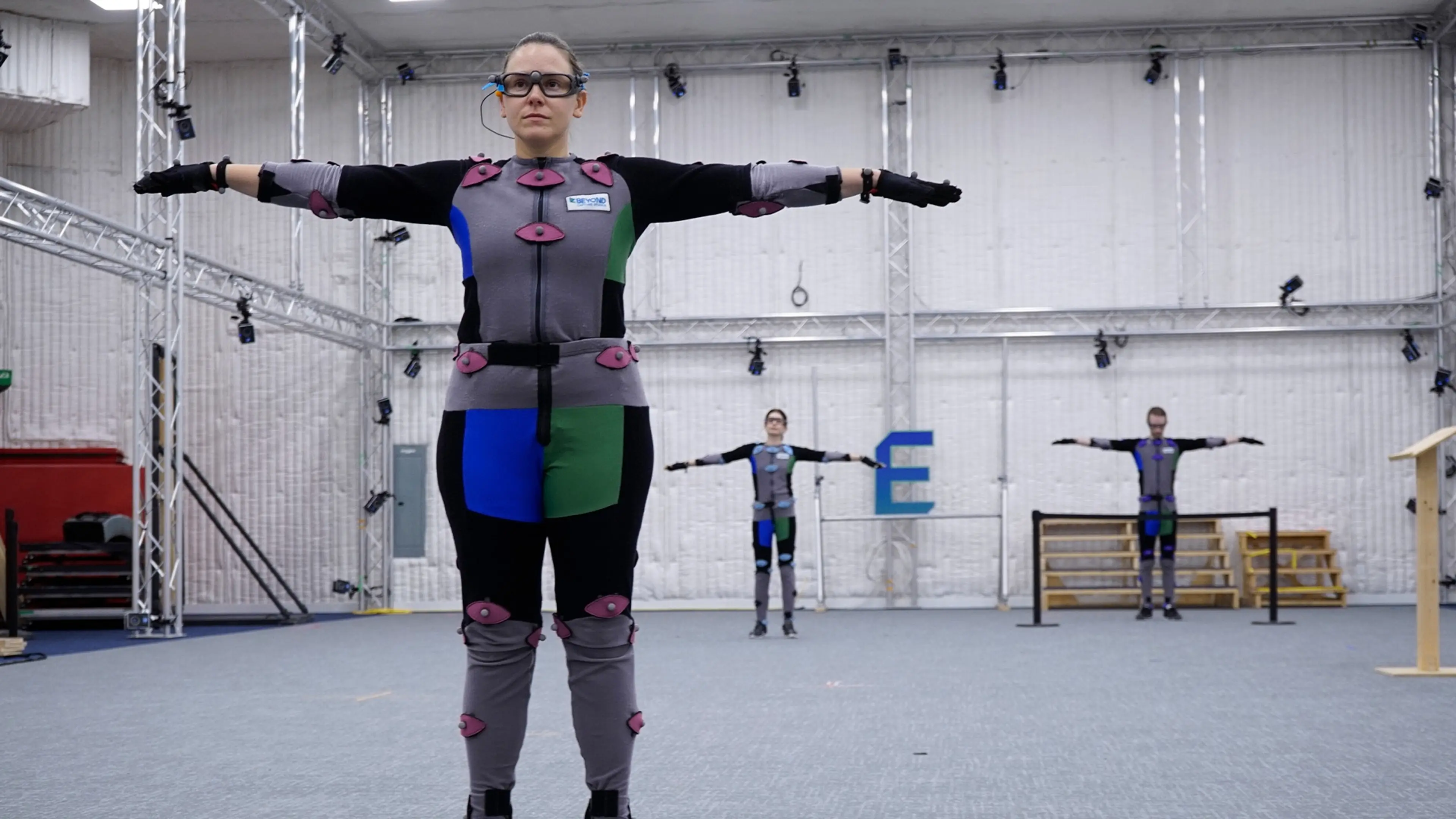 Three people in motion capture suits with sensors stand with arms outstretched in a large studio space.