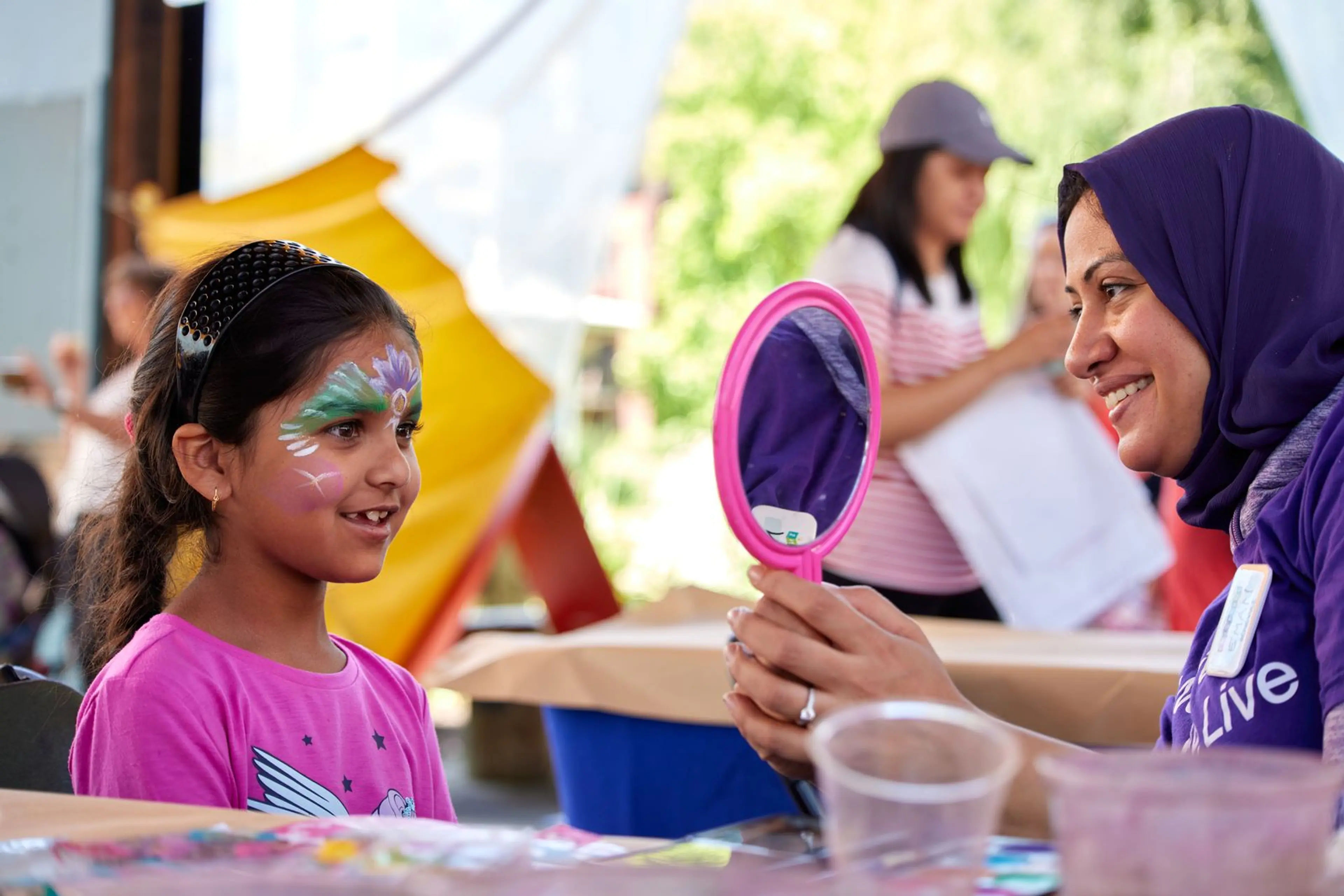 A child with face paint looks at their reflection in a handheld mirror held by a smiling person wearing a purple hijab and shirt.