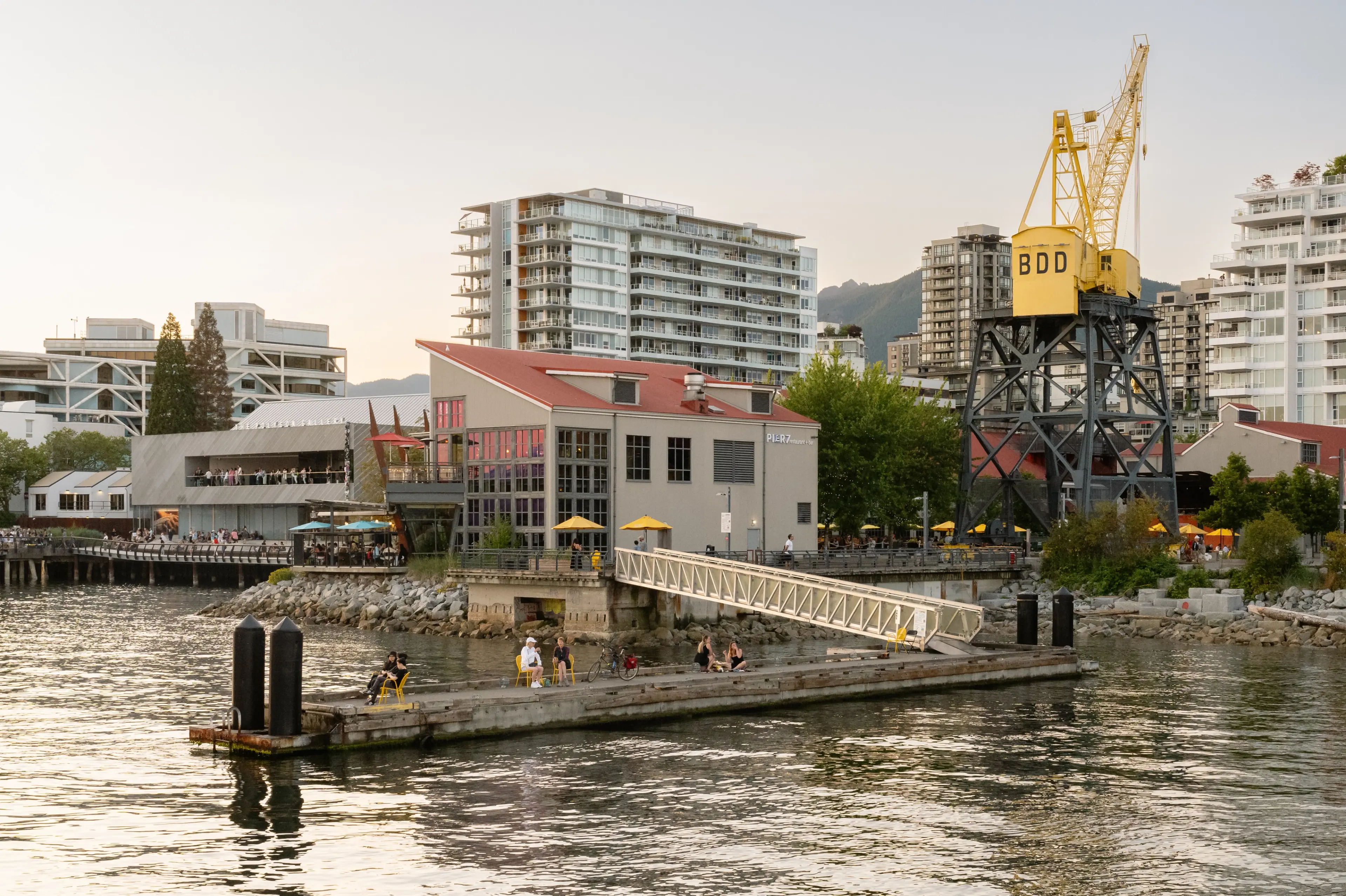 Waterfront scene with a dock, people sitting and walking, a yellow crane labeled BDD, and modern buildings in the background.