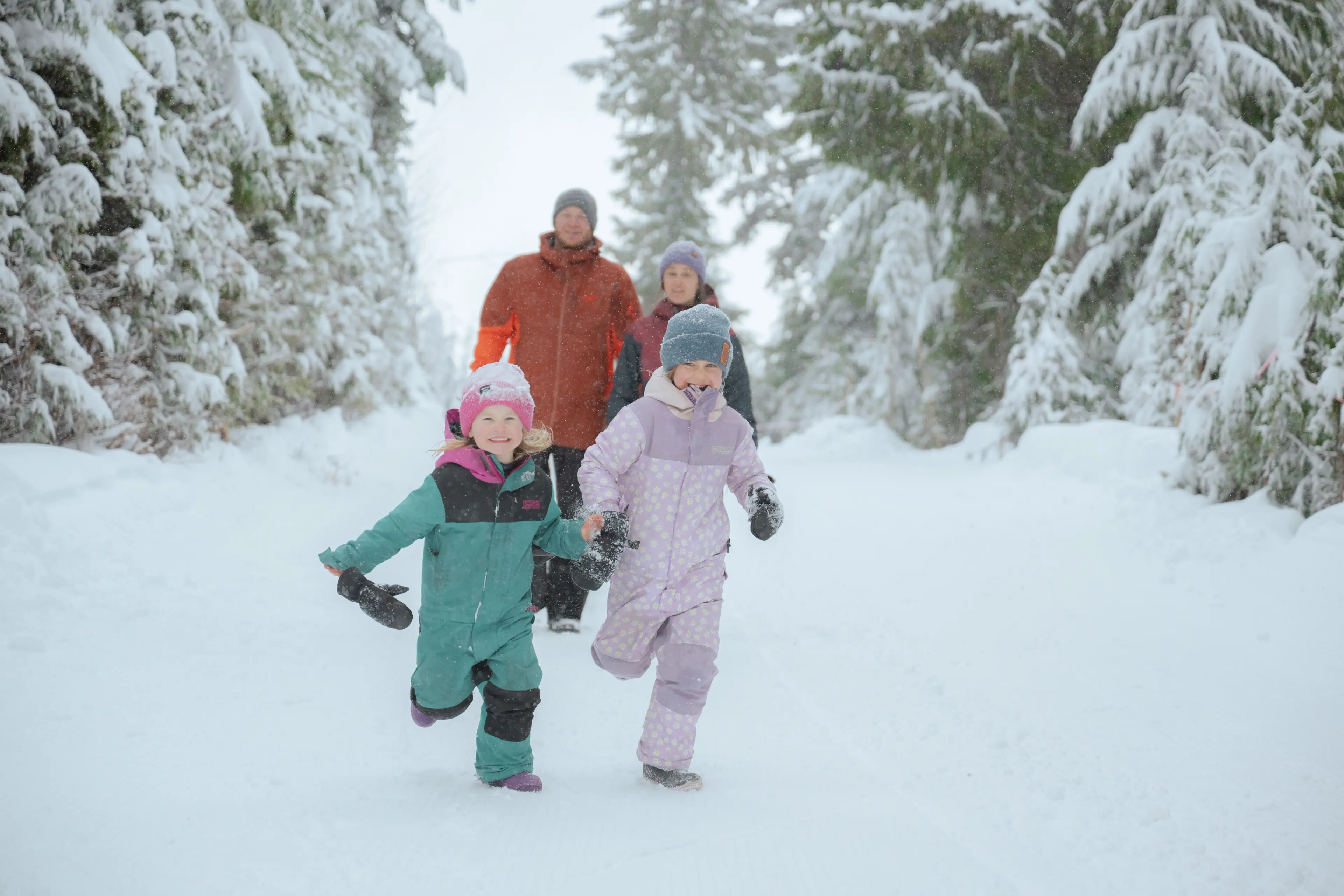 Two children in colorful snowsuits running on a snowy path with two adults walking behind them among snow-covered trees.