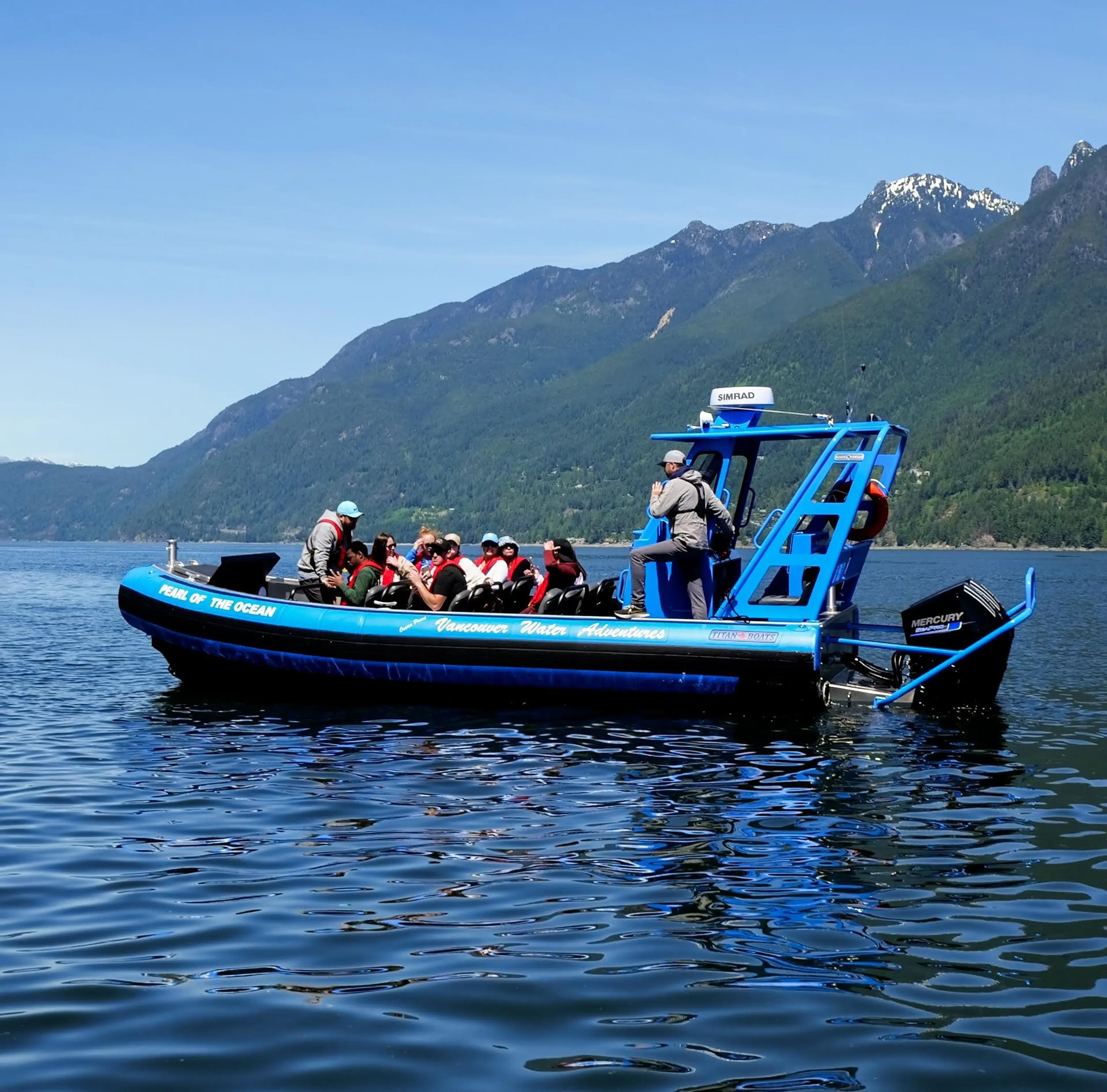 Blue tour boat with passengers on calm water near forested mountains under clear sky.