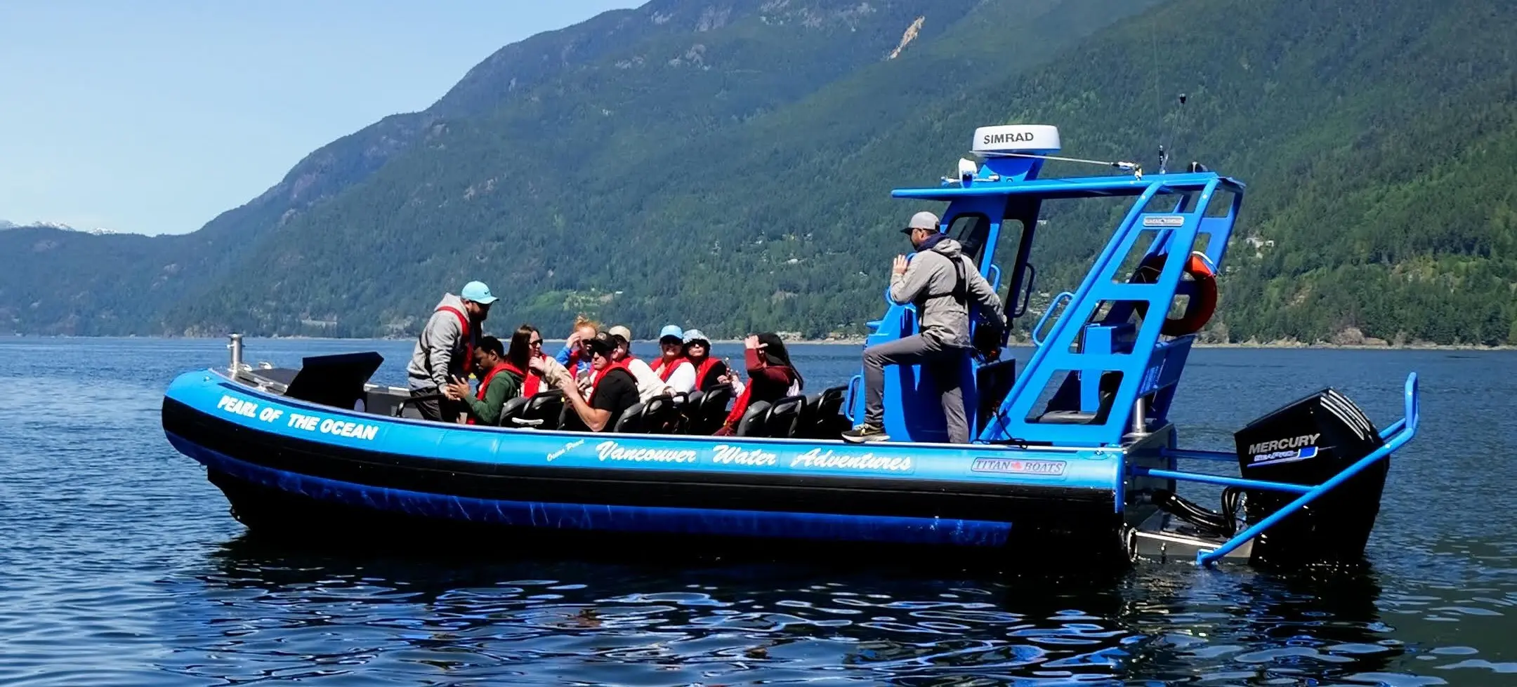 Blue tour boat with passengers on calm water near forested mountains under clear sky.