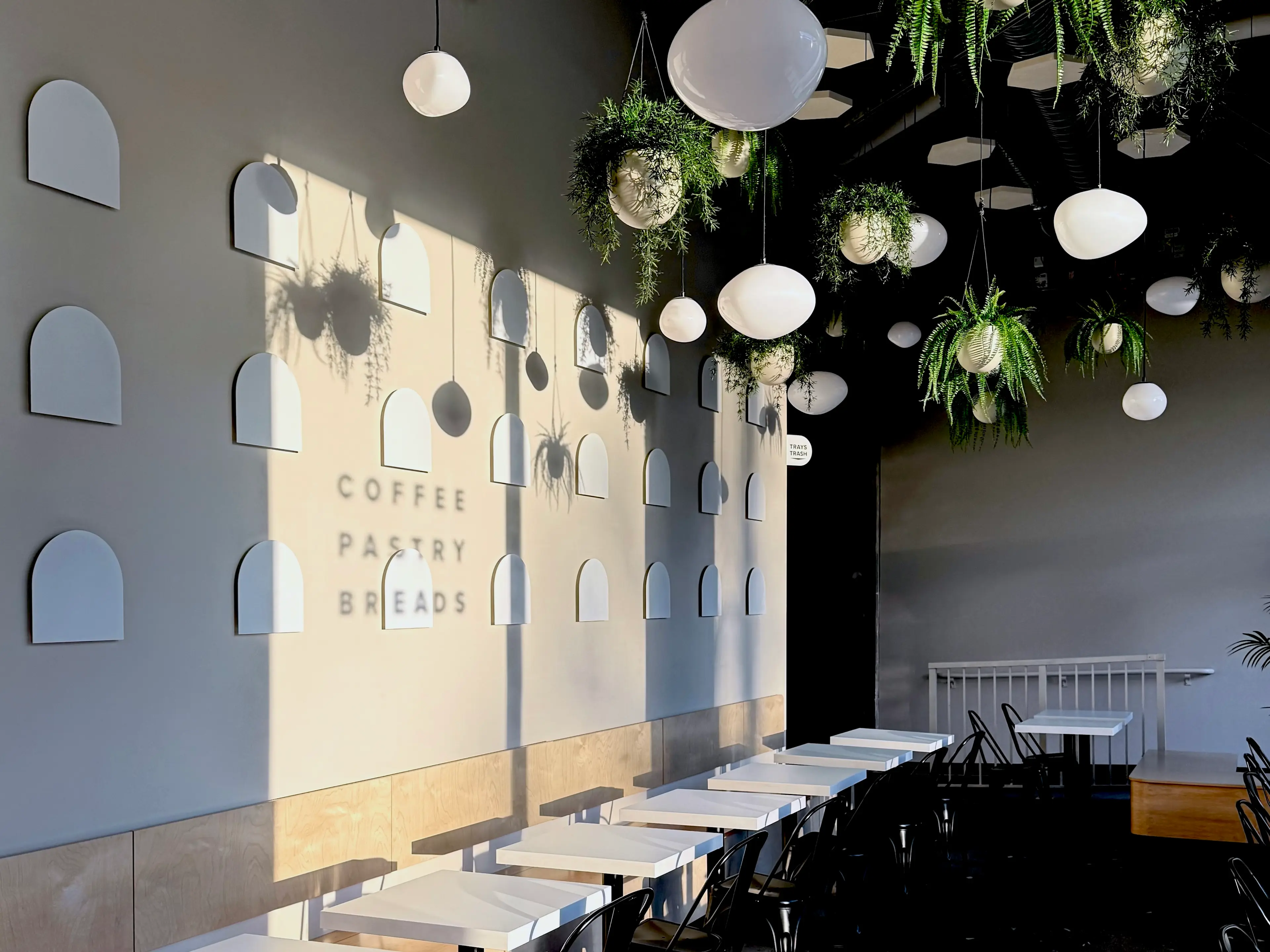 Modern cafe interior with white tables, black chairs, hanging plants, and wall decor casting shadows with words coffee, pastry, breads.
