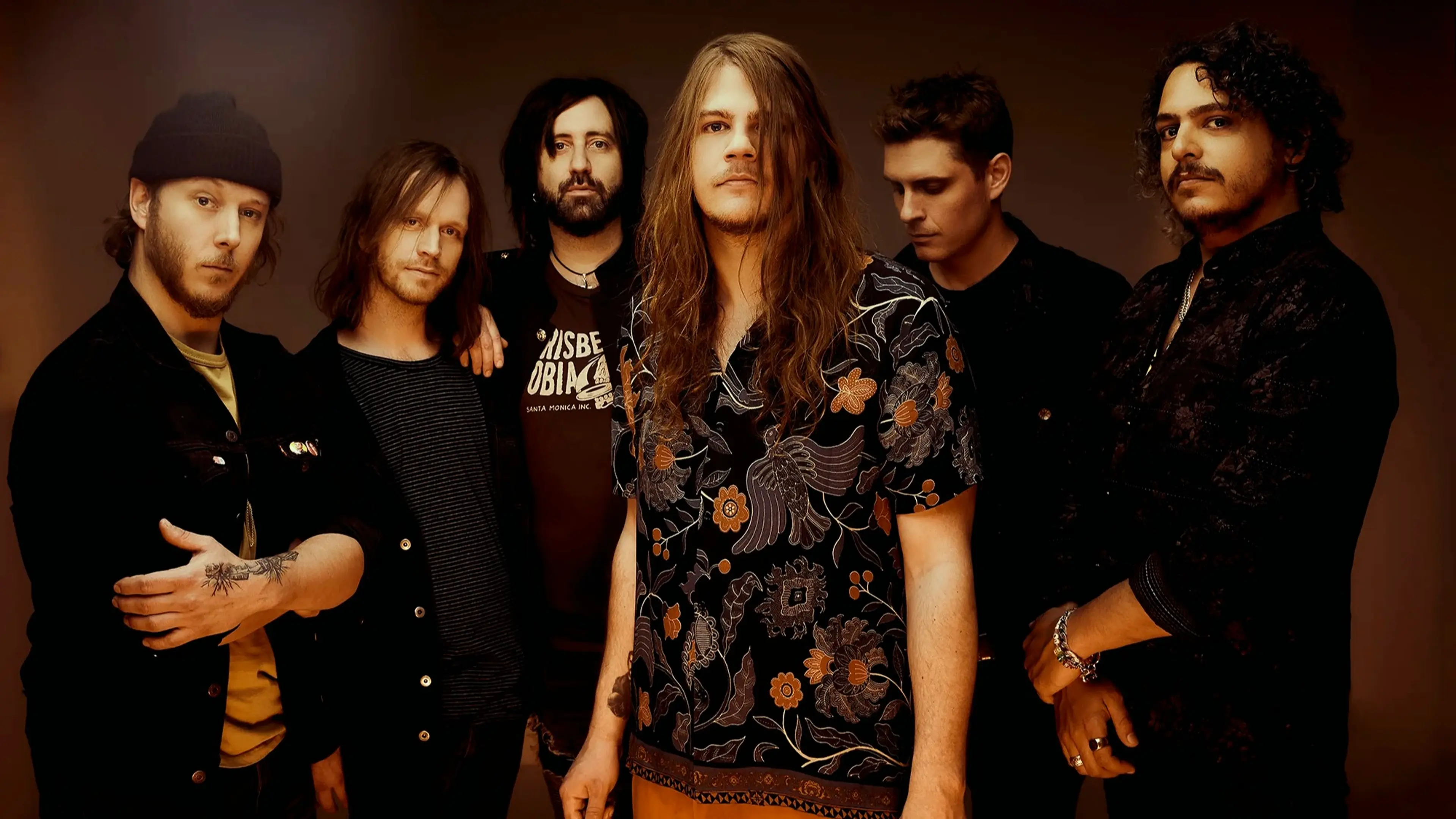 Six people standing in a row against a brown background, wearing casual dark clothing, one with long hair and a floral shirt in the center.
