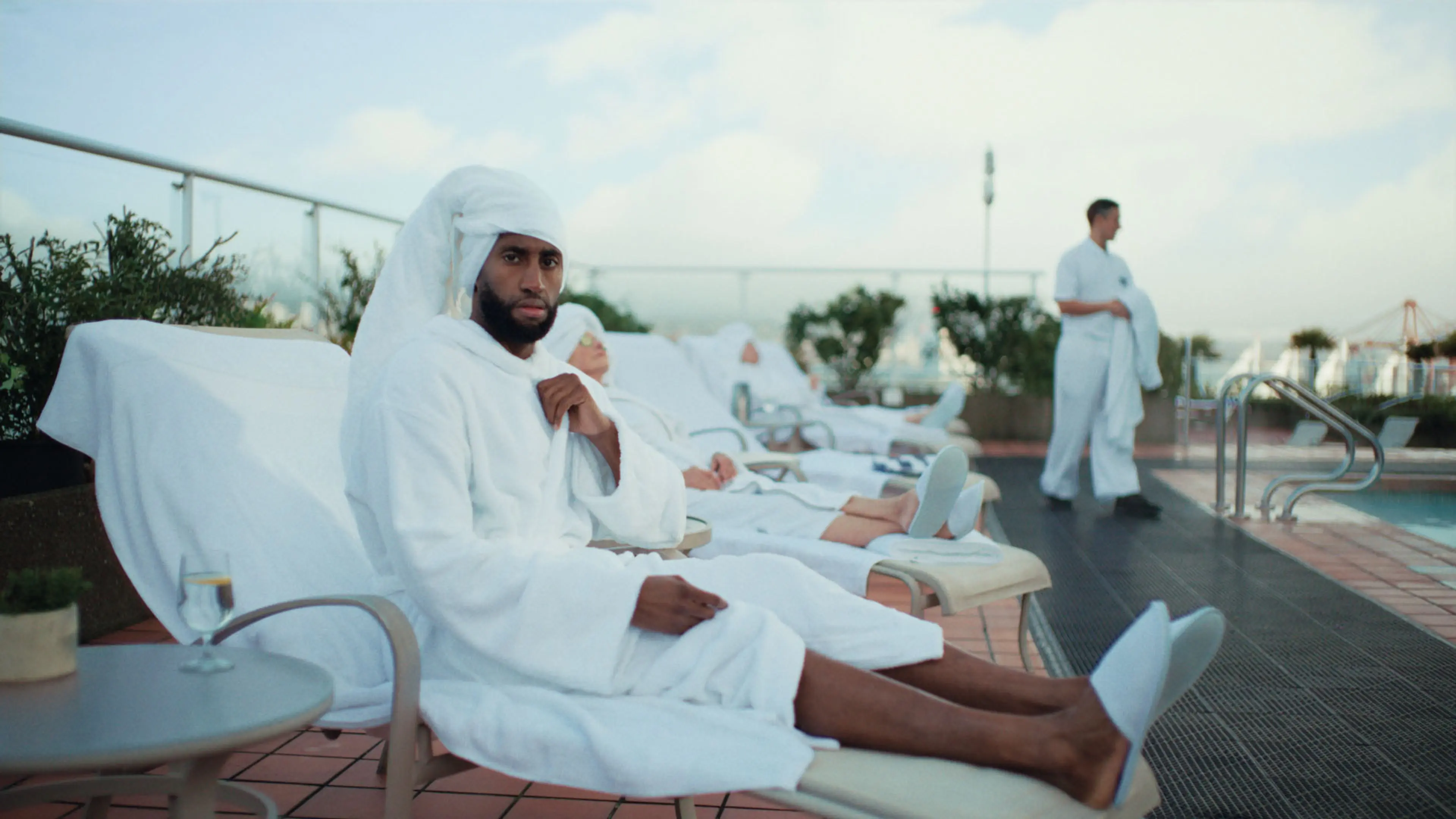 Man in white bathrobe and towel turban sitting on a lounge chair by the pool with others relaxing in the background.