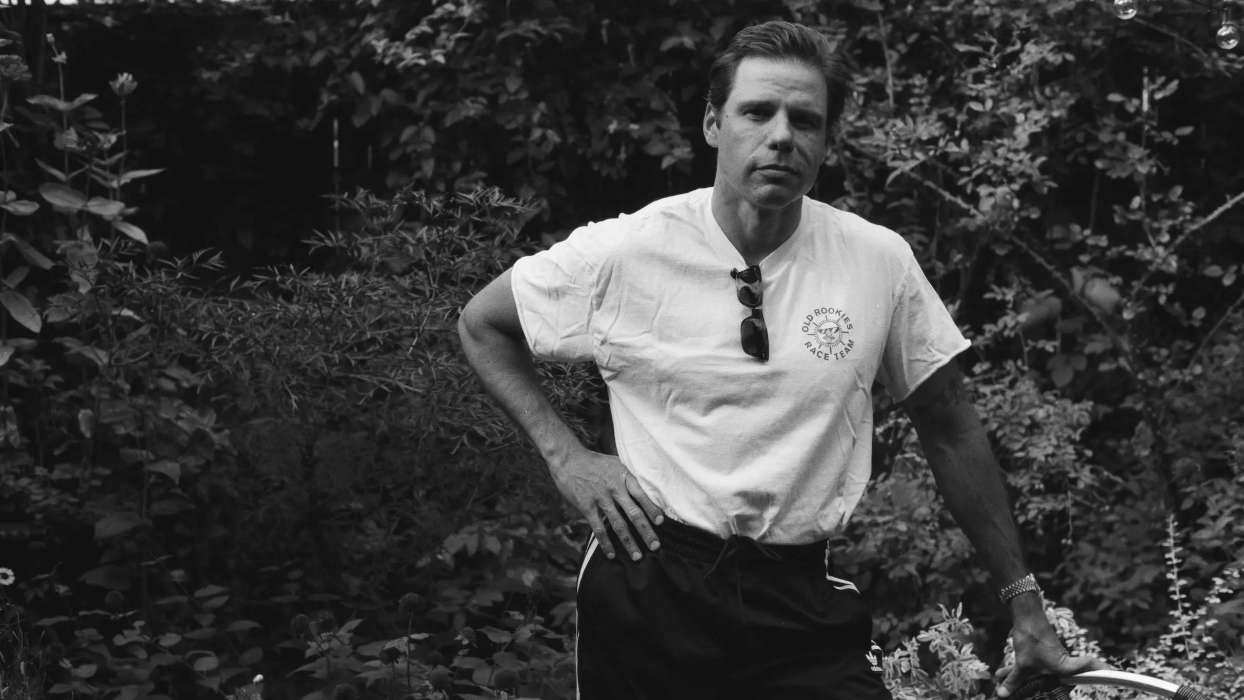 Person in a white t-shirt and dark pants standing outdoors with one hand on hip and the other resting on a bicycle handlebar.