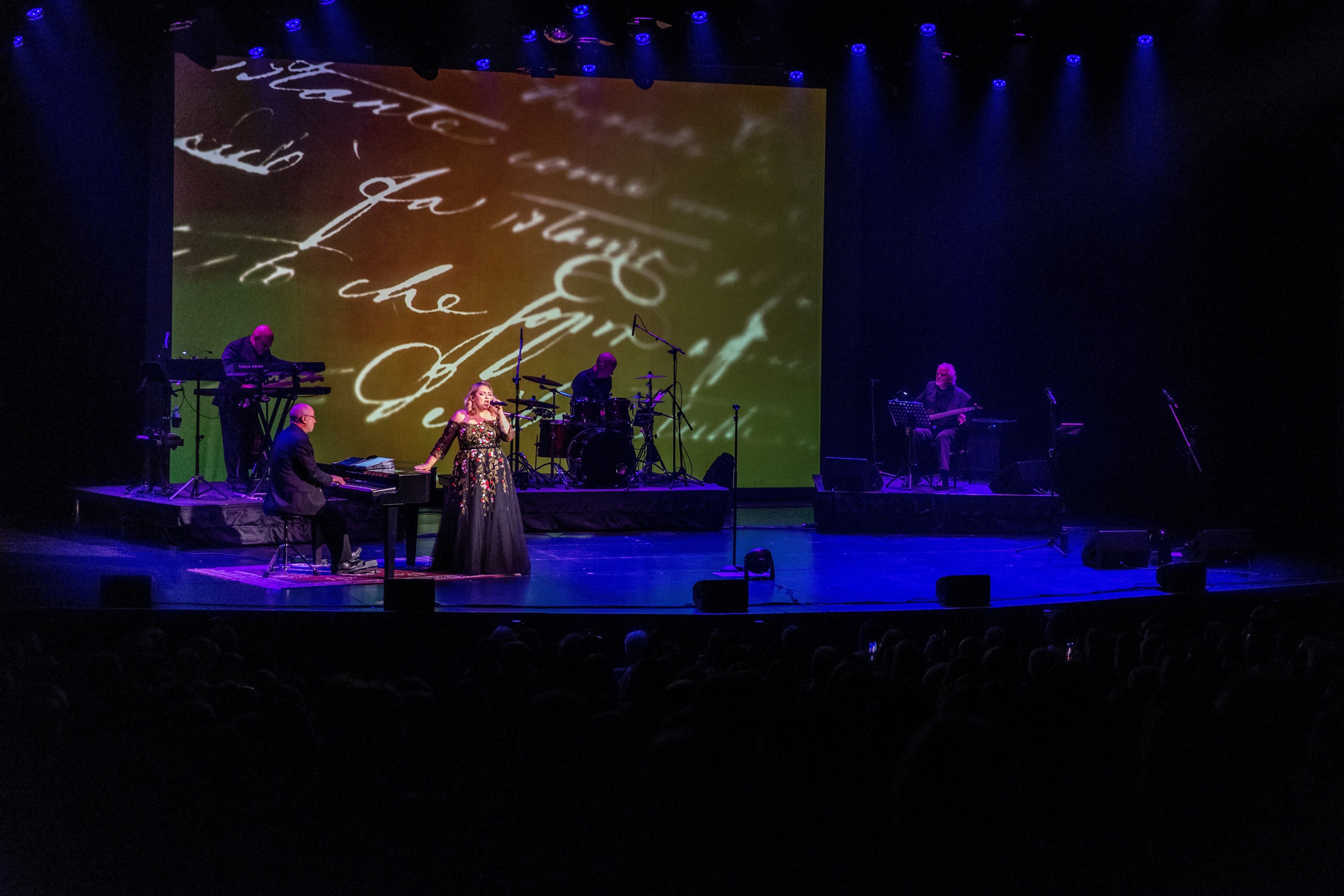 A singer in a black floral dress performs on stage with a pianist, drummer, and keyboardist, with handwritten text projected behind.