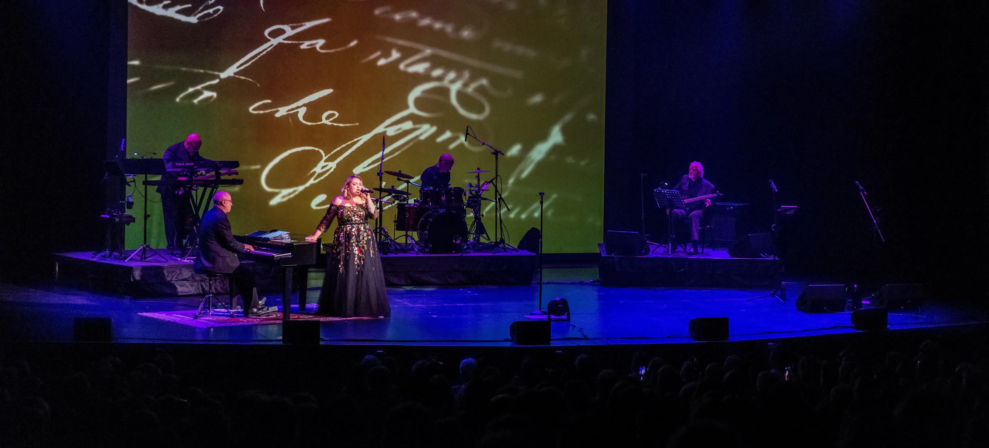 A singer in a black floral dress performs on stage with a pianist, drummer, and keyboardist, with handwritten text projected behind.