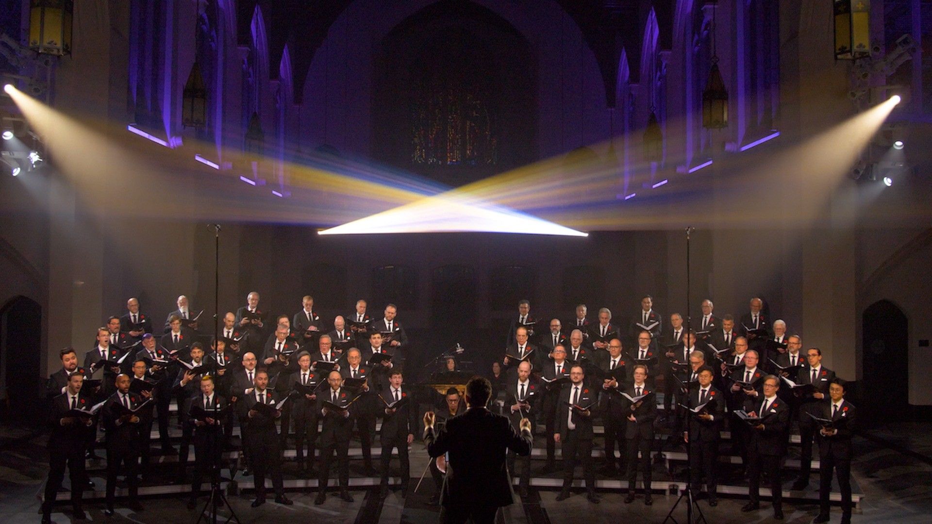 Large choir of men in black suits singing in a church with a conductor in front and stage lights above.