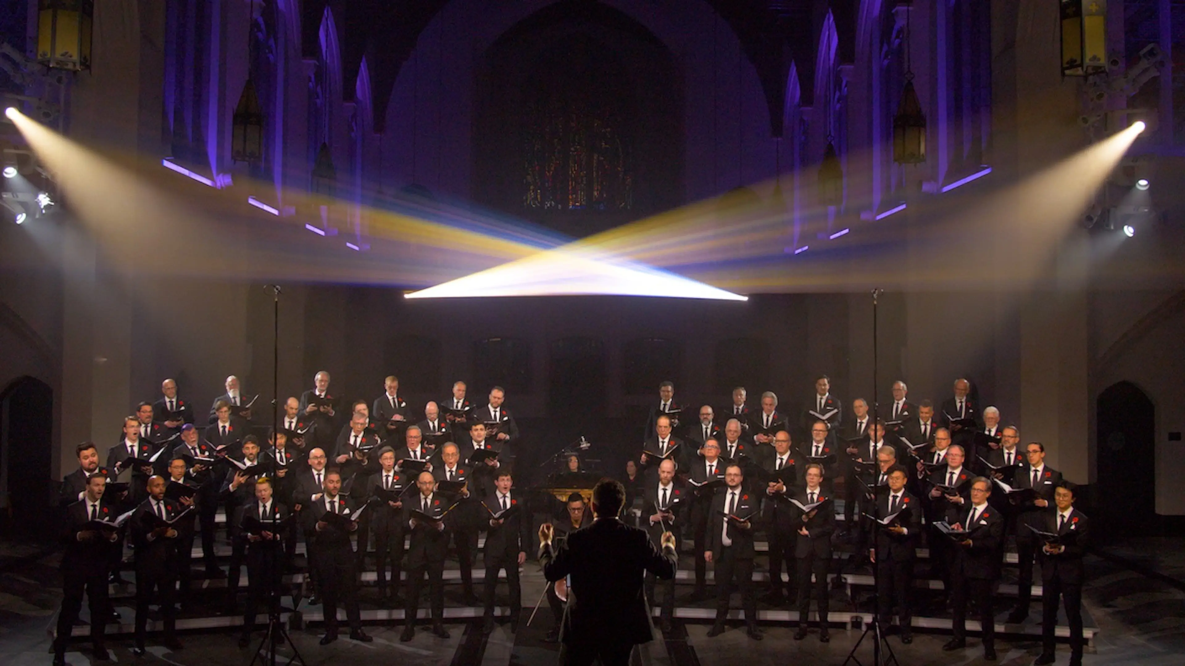 Large choir of men in black suits singing in a church with a conductor in front and stage lights above.