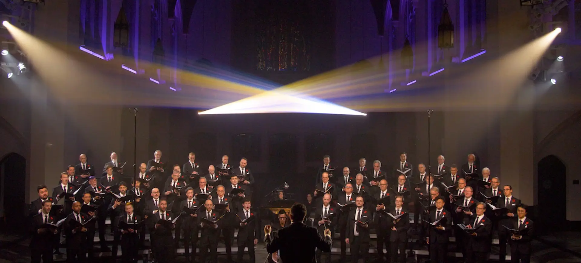 Large choir of men in black suits singing in a church with a conductor in front and stage lights above.