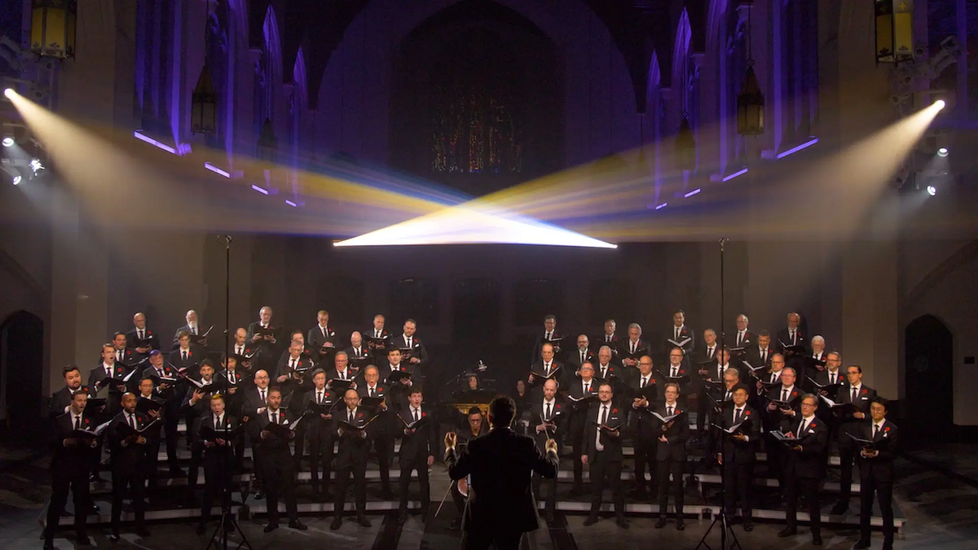 Large choir of men in black suits singing in a church with a conductor in front and stage lights above.