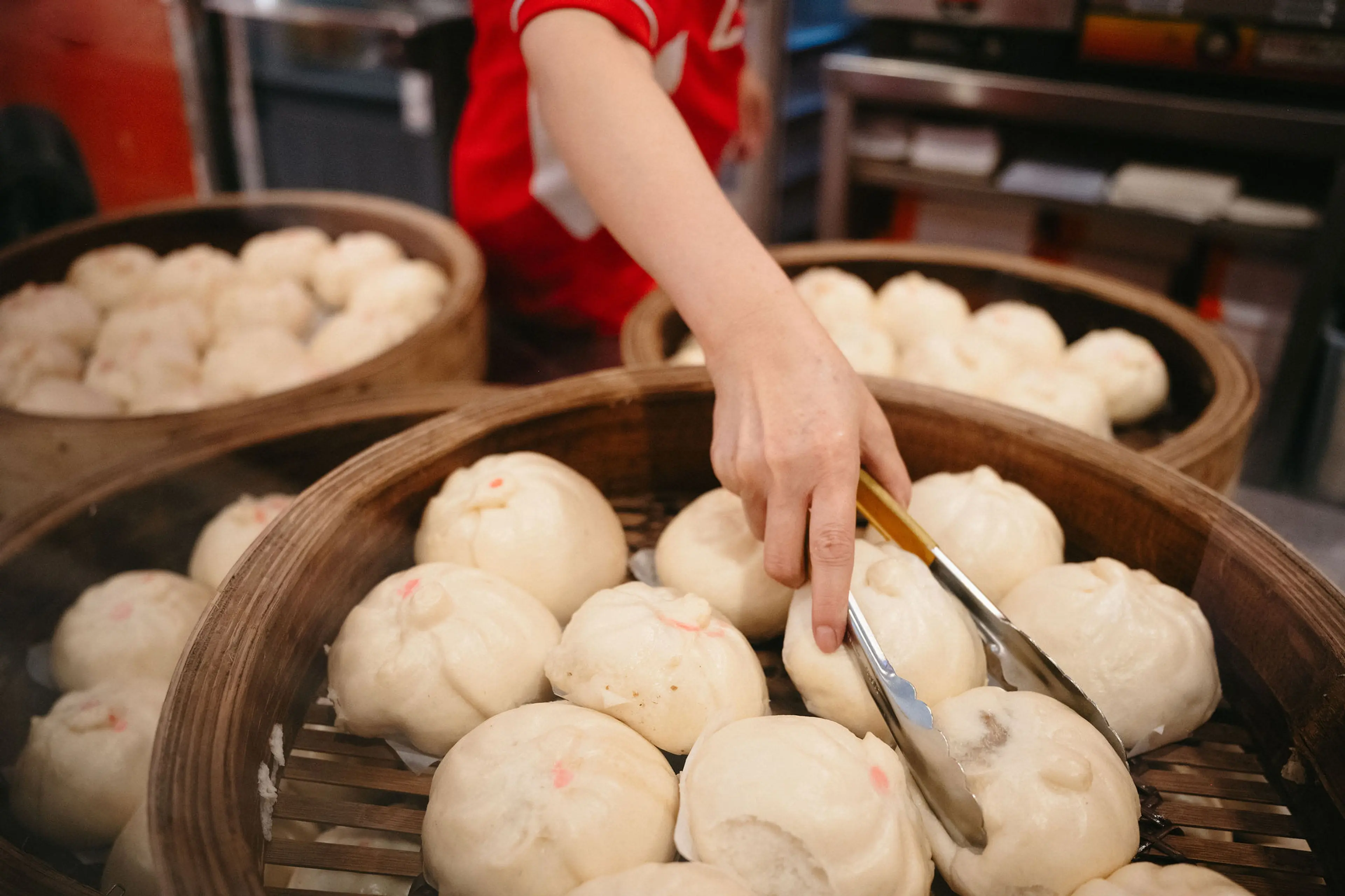 A person using tongs to pick steamed buns from a bamboo steamer basket filled with buns.