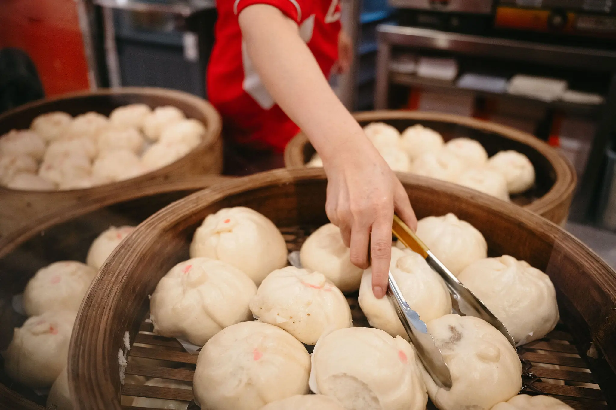 A person using tongs to pick steamed buns from a bamboo steamer basket filled with buns.