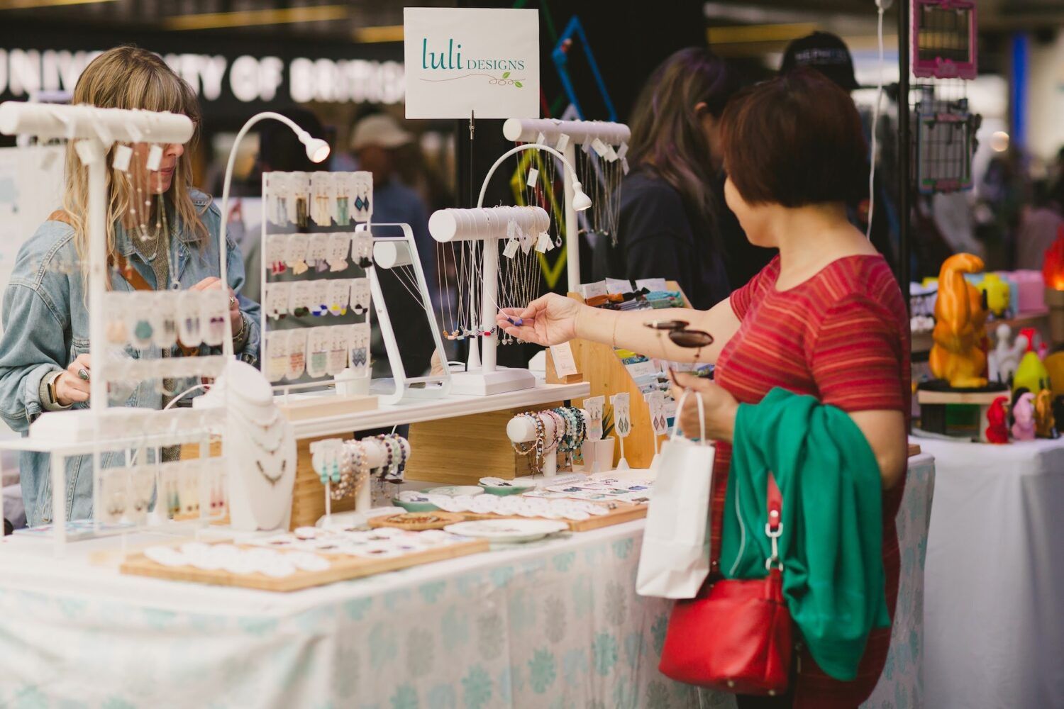 A person in a red striped shirt browsing jewelry at a market stall labeled Luli Designs, with another person behind the counter.