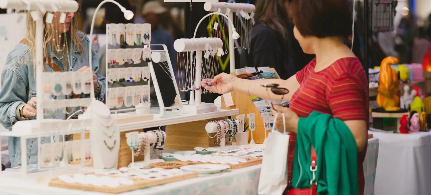 A person in a red striped shirt browsing jewelry at a market stall labeled Luli Designs, with another person behind the counter.