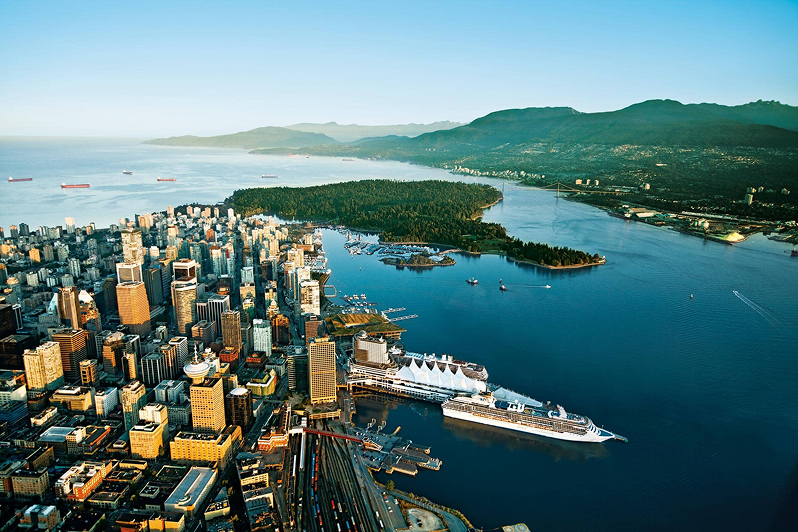 Aerial view of a coastal city with tall buildings, a large cruise ship docked, a forested peninsula, and mountains in the background.