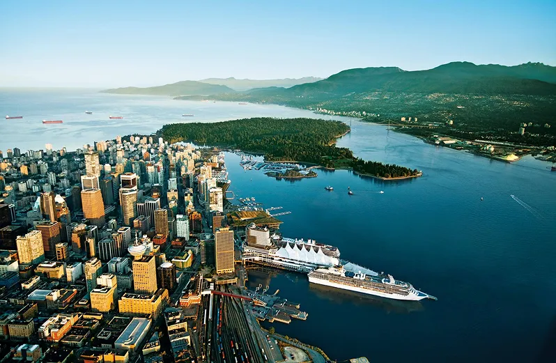 Aerial view of a coastal city with tall buildings, a large cruise ship docked, a forested peninsula, and mountains in the background.