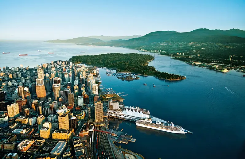Aerial view of a coastal city with tall buildings, a large cruise ship docked, a forested peninsula, and mountains in the background.