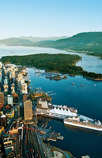 Aerial view of a coastal city with tall buildings, a large cruise ship docked, a forested peninsula, and mountains in the background.