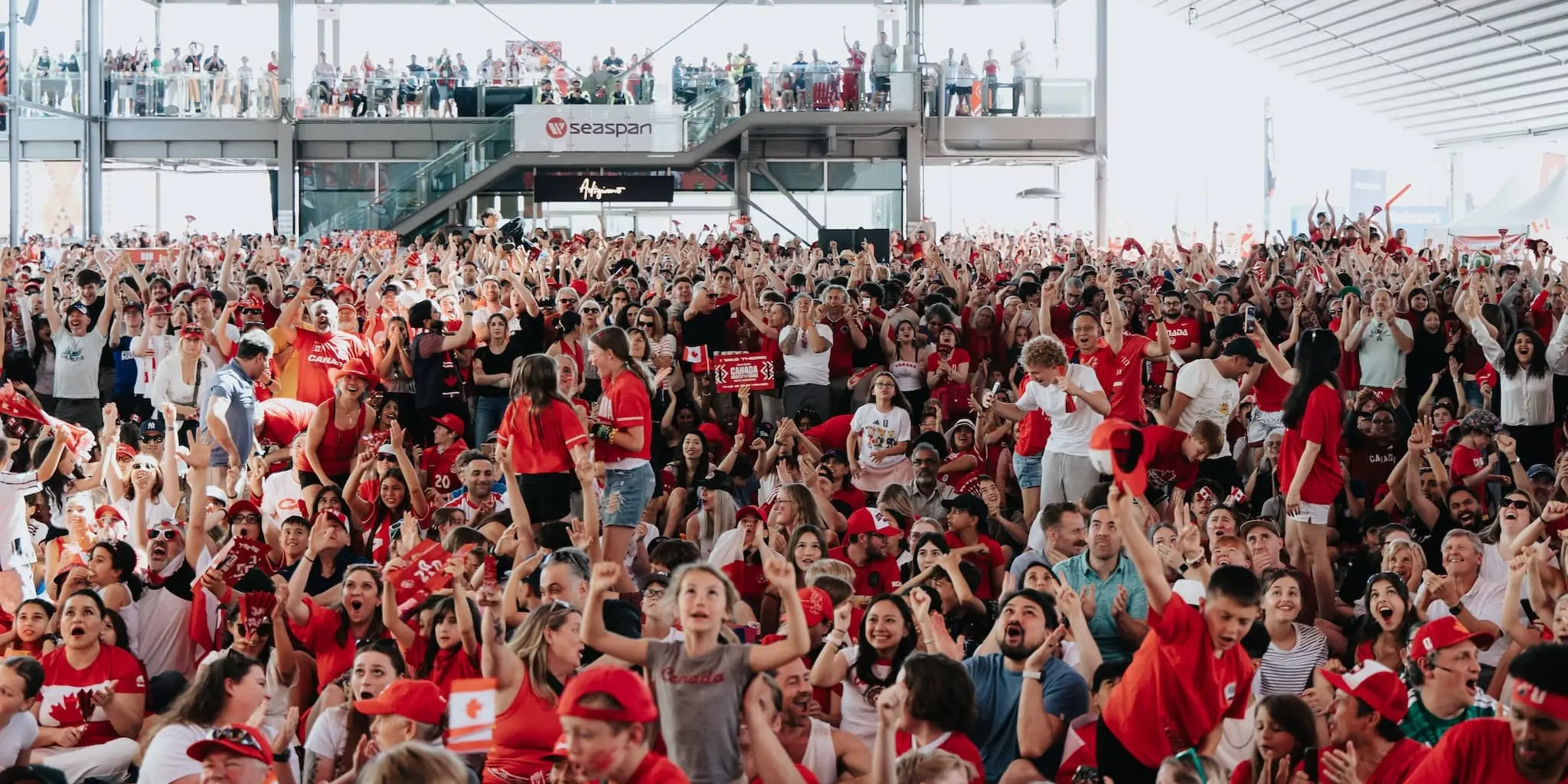 Large crowd of people wearing red and white, many holding phones, one person in white shirt with red maple leaf raising hand.
