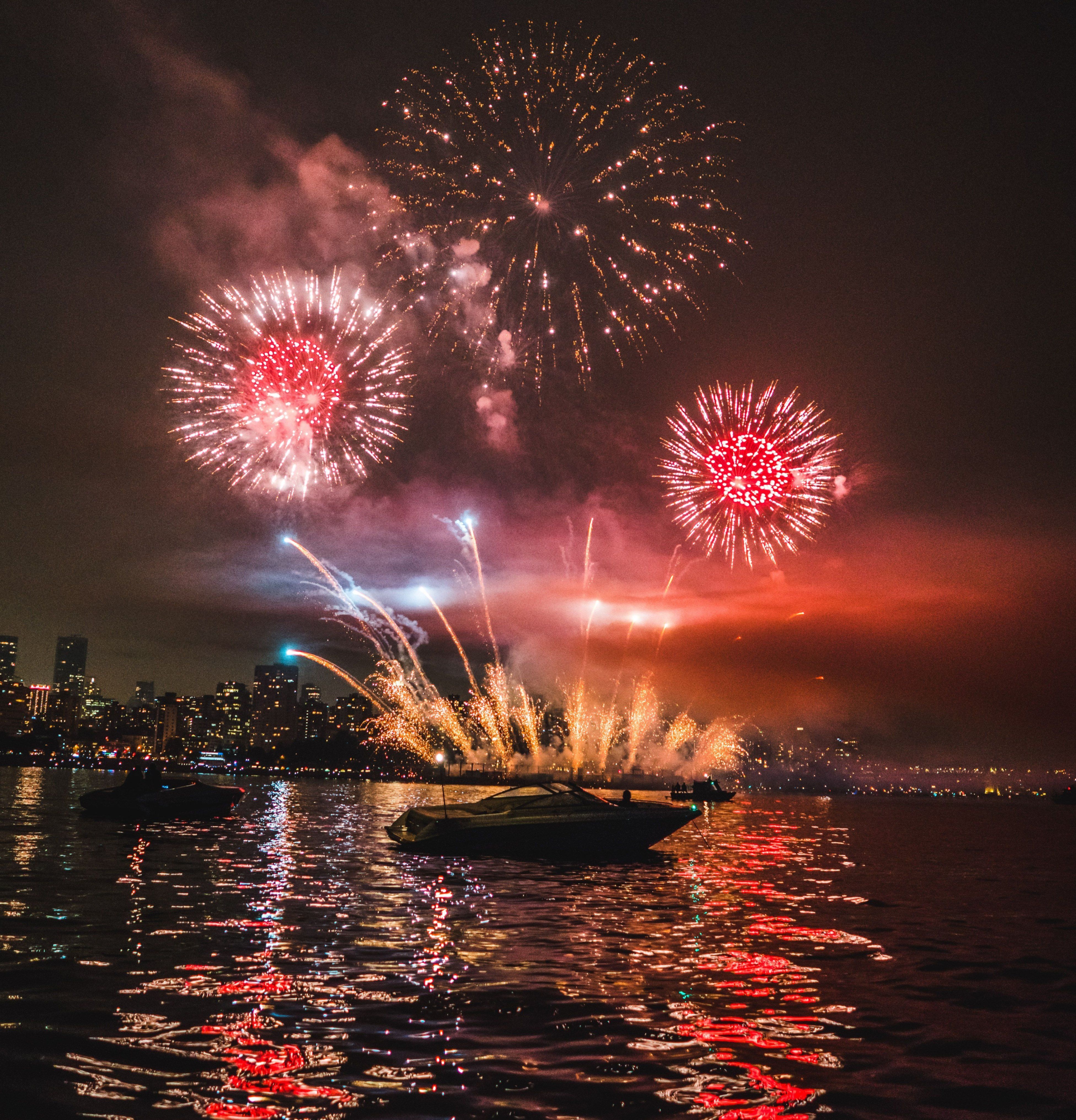 Fireworks exploding over a city skyline at night, reflecting on water with boats in the foreground.