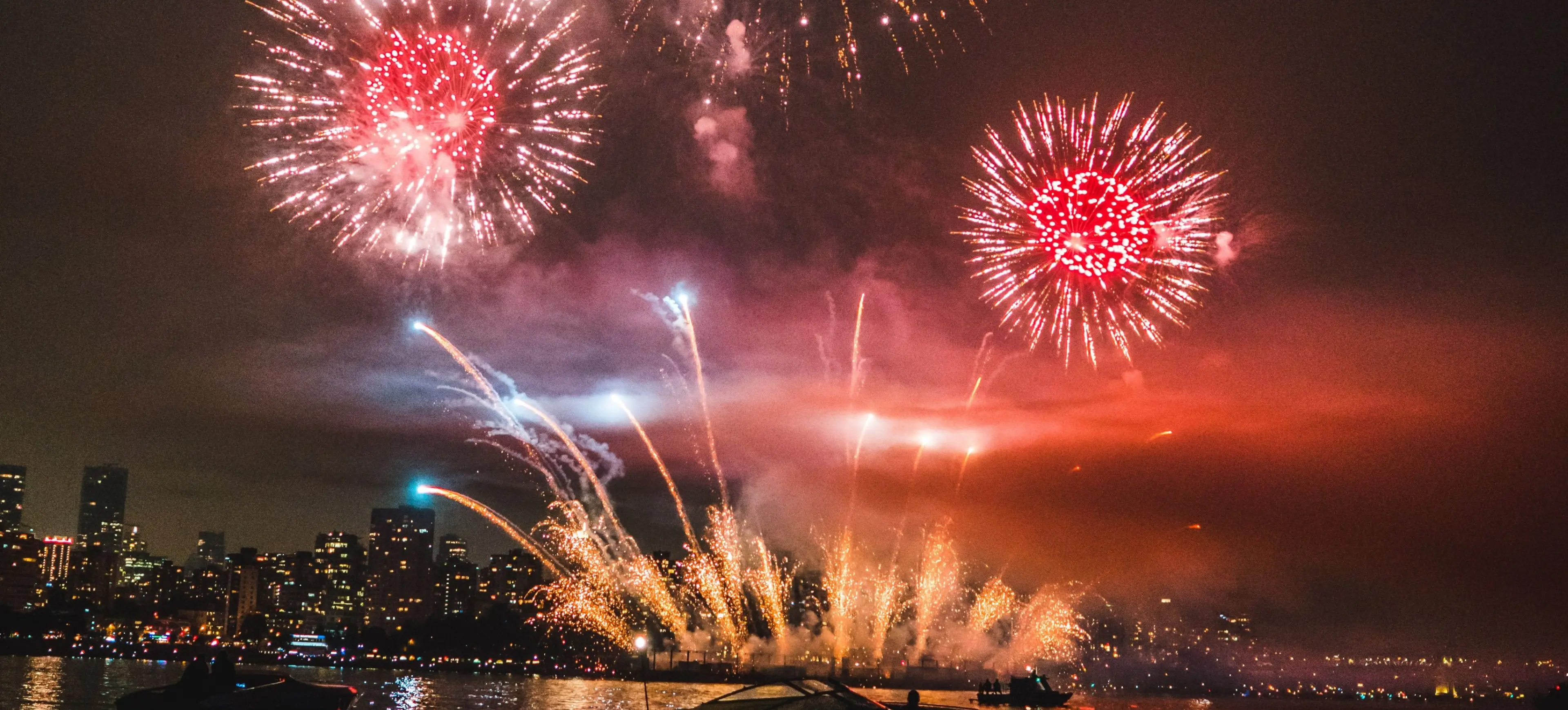 Fireworks exploding over a city skyline at night, reflecting on water with boats in the foreground.