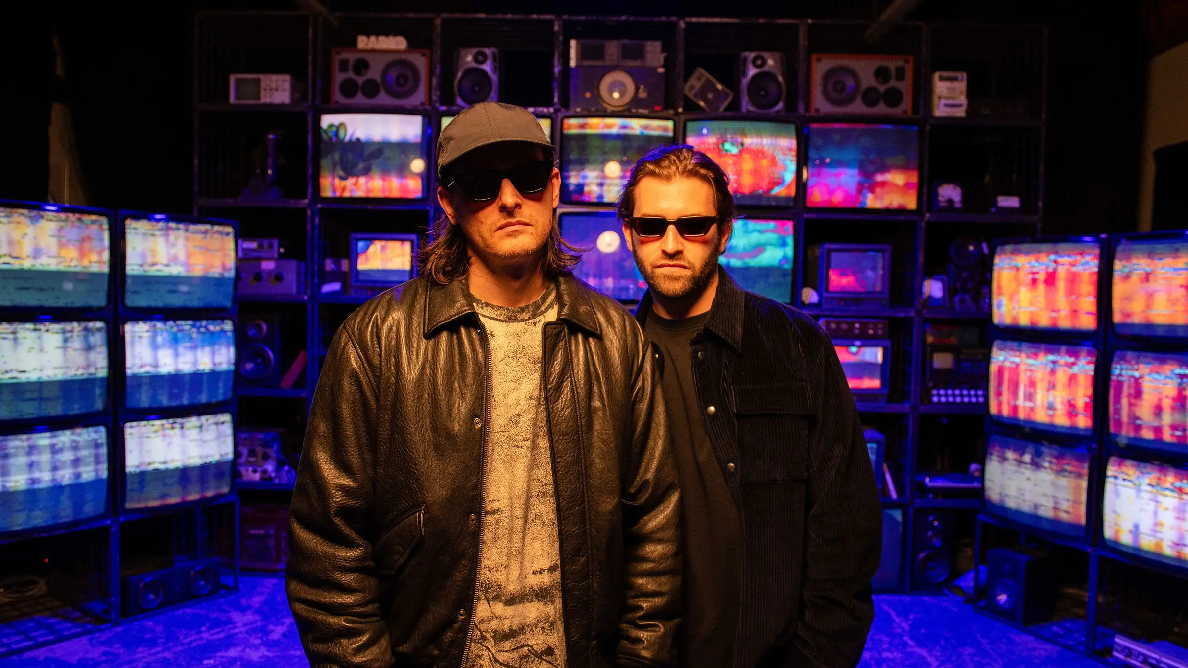Two people wearing sunglasses and dark jackets standing in front of a wall of colorful, glitching TV screens.