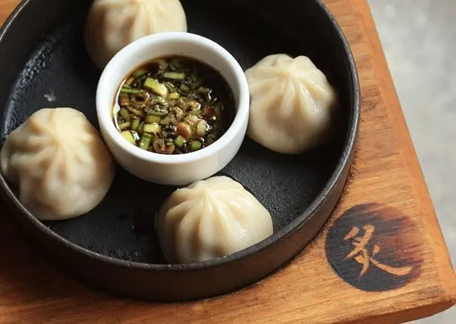 Four steamed dumplings arranged around a small bowl of dipping sauce with chopped green onions in a black round dish on a wooden board.