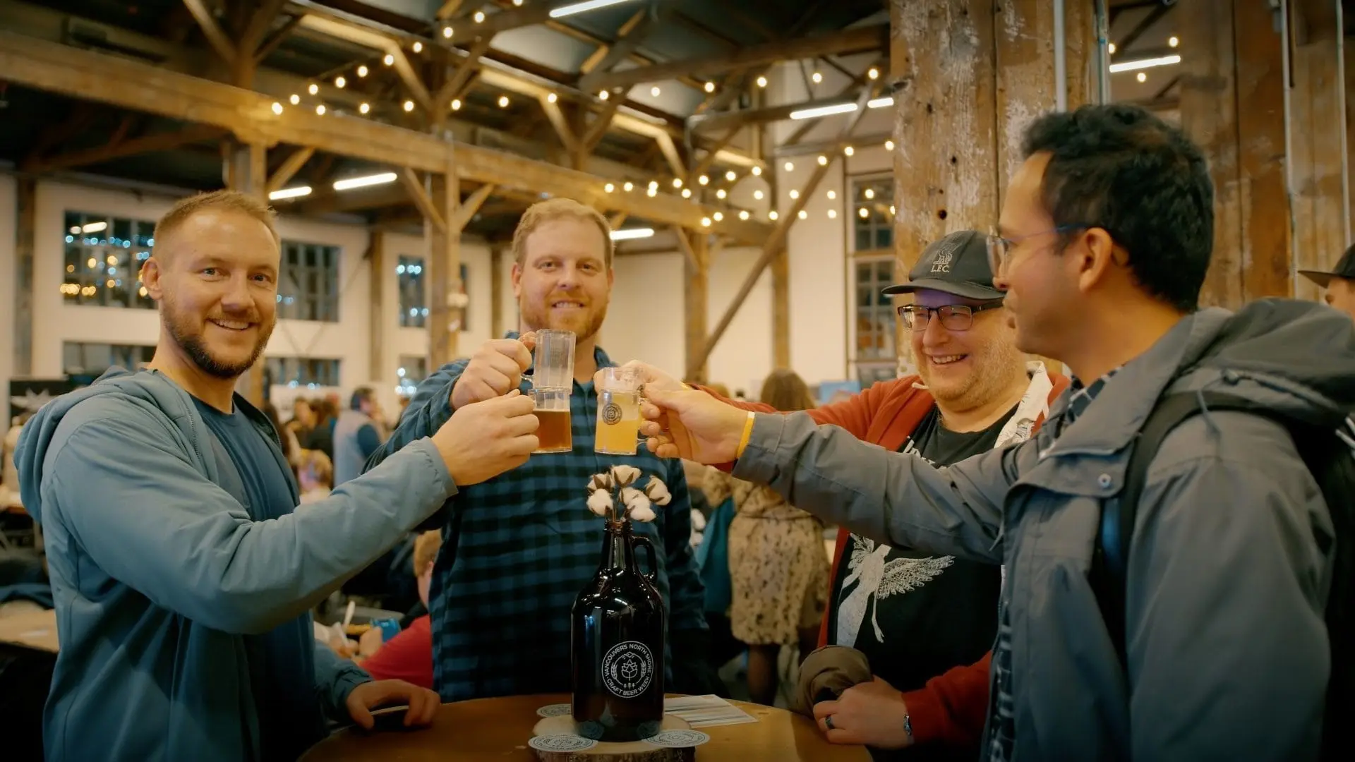 Four people clinking glasses in a warmly lit indoor space with string lights and wooden beams.