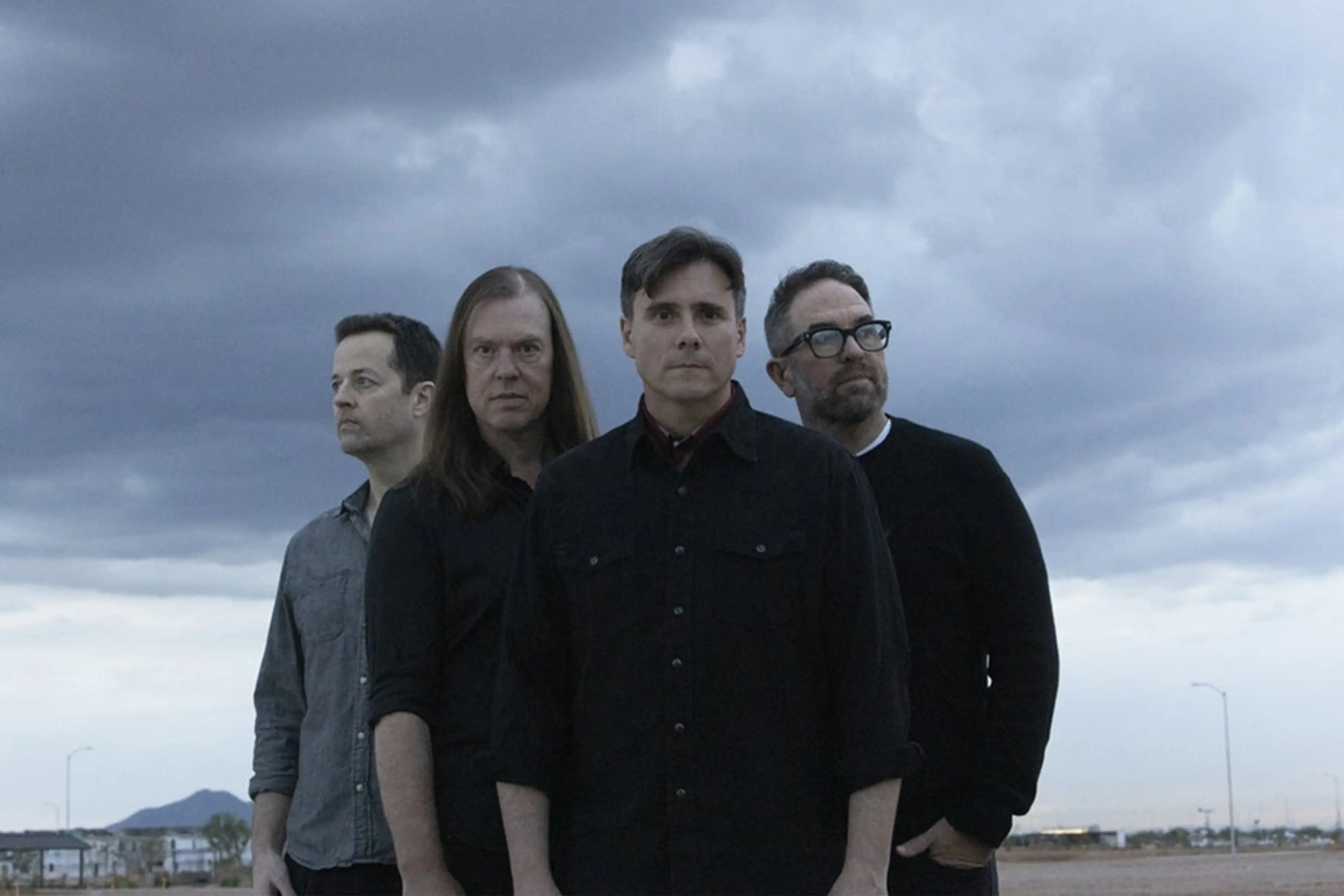 Four people standing outdoors under a cloudy sky, looking in different directions, wearing dark and gray shirts.