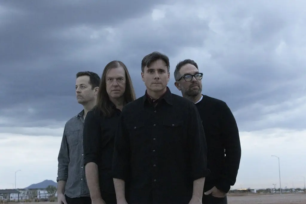 Four people standing outdoors under a cloudy sky, looking in different directions, wearing dark and gray shirts.