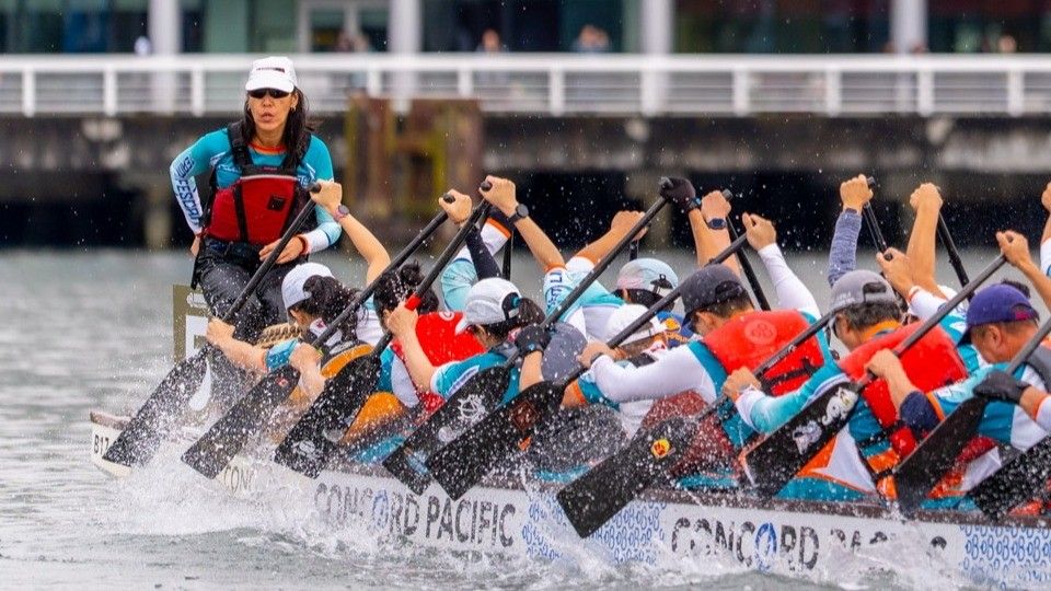 A team paddling vigorously in a dragon boat race on water, led by a person standing at the front.