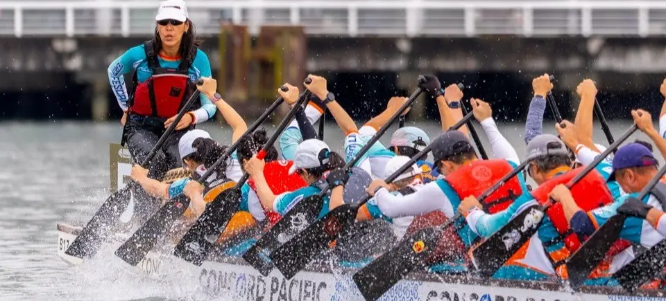 A team paddling vigorously in a dragon boat race on water, led by a person standing at the front.