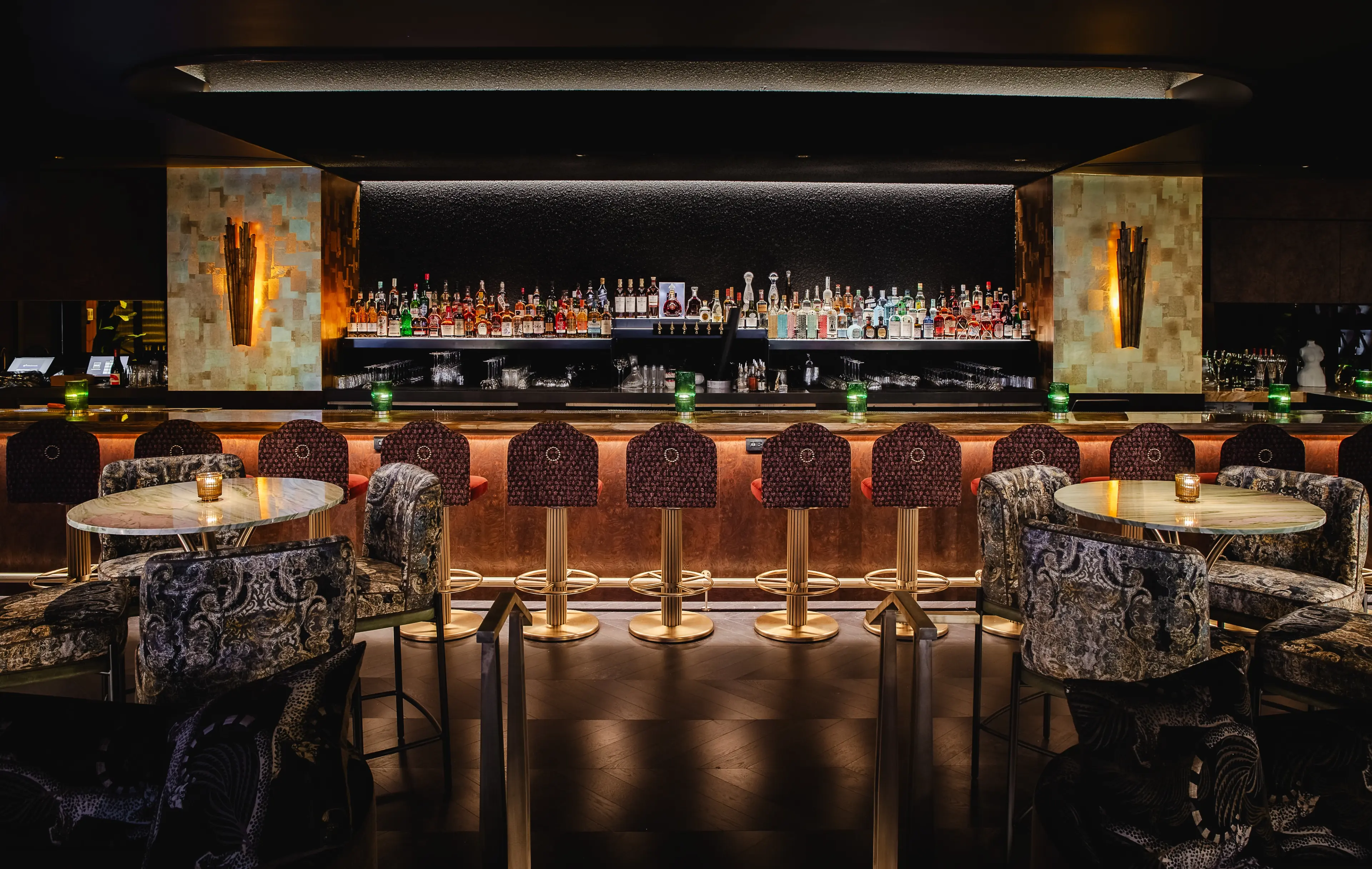 The interior of Prophecy Bar at the Rosewood Hotel Georgia. The long, dark bar has rows of top shelf alcohol behind it and upholstered bar stools in front of it.