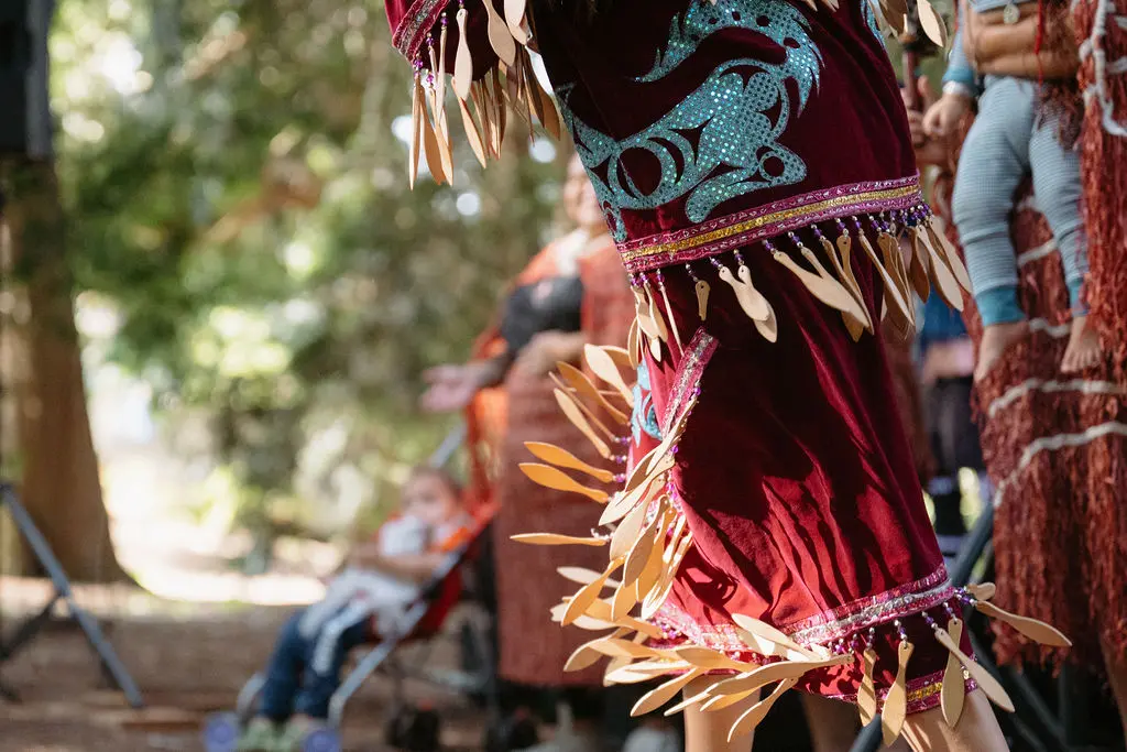 Close-up of a person wearing a maroon traditional outfit with blue patterns and wooden decorations, dancing outdoors.