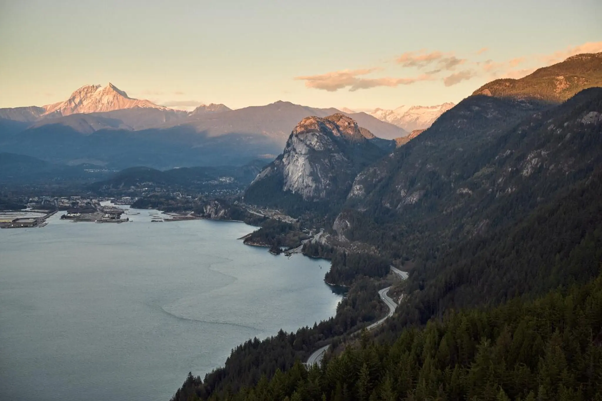 Mountain landscape with a winding road along a forested shoreline and snow-capped peaks in the distance at sunset.