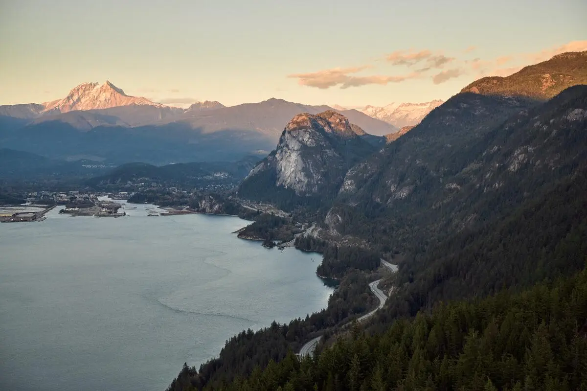 Mountain landscape with a winding road along a forested shoreline and snow-capped peaks in the distance at sunset.