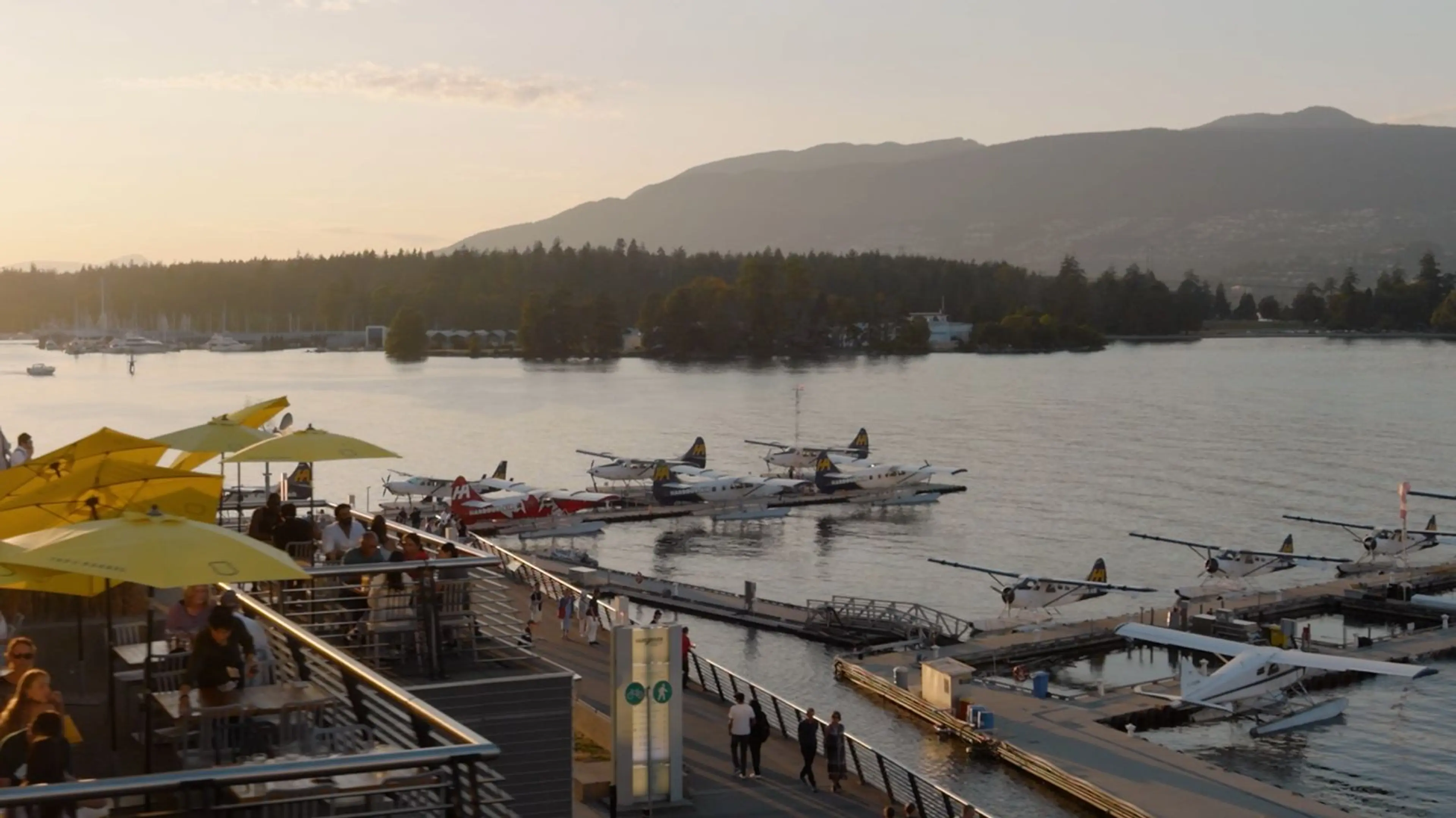 Seaplanes docked at a marina with people sitting under yellow umbrellas on a terrace overlooking the water at sunset.