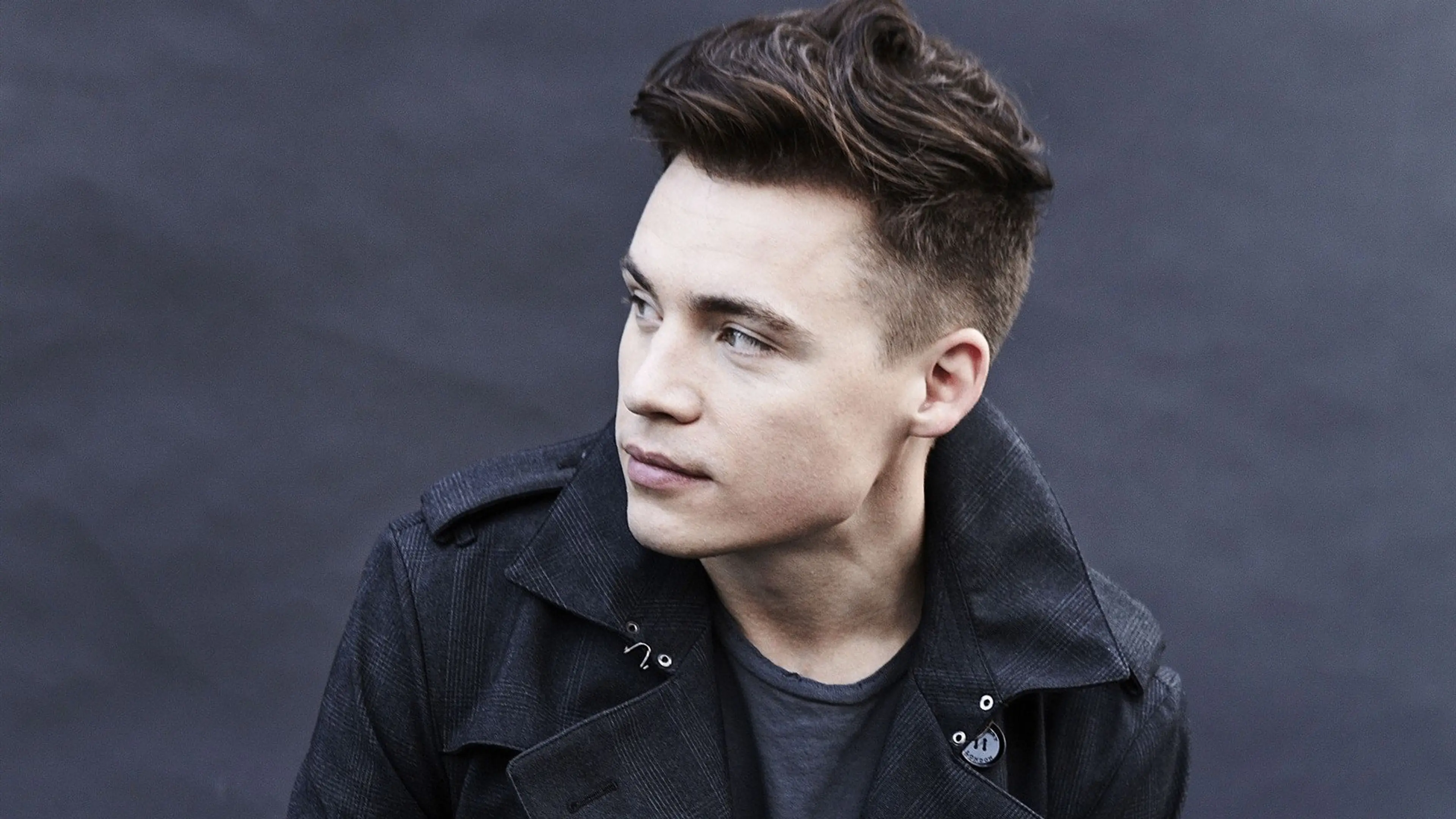 Person with short styled hair wearing a dark jacket looking to the side against a dark background.
