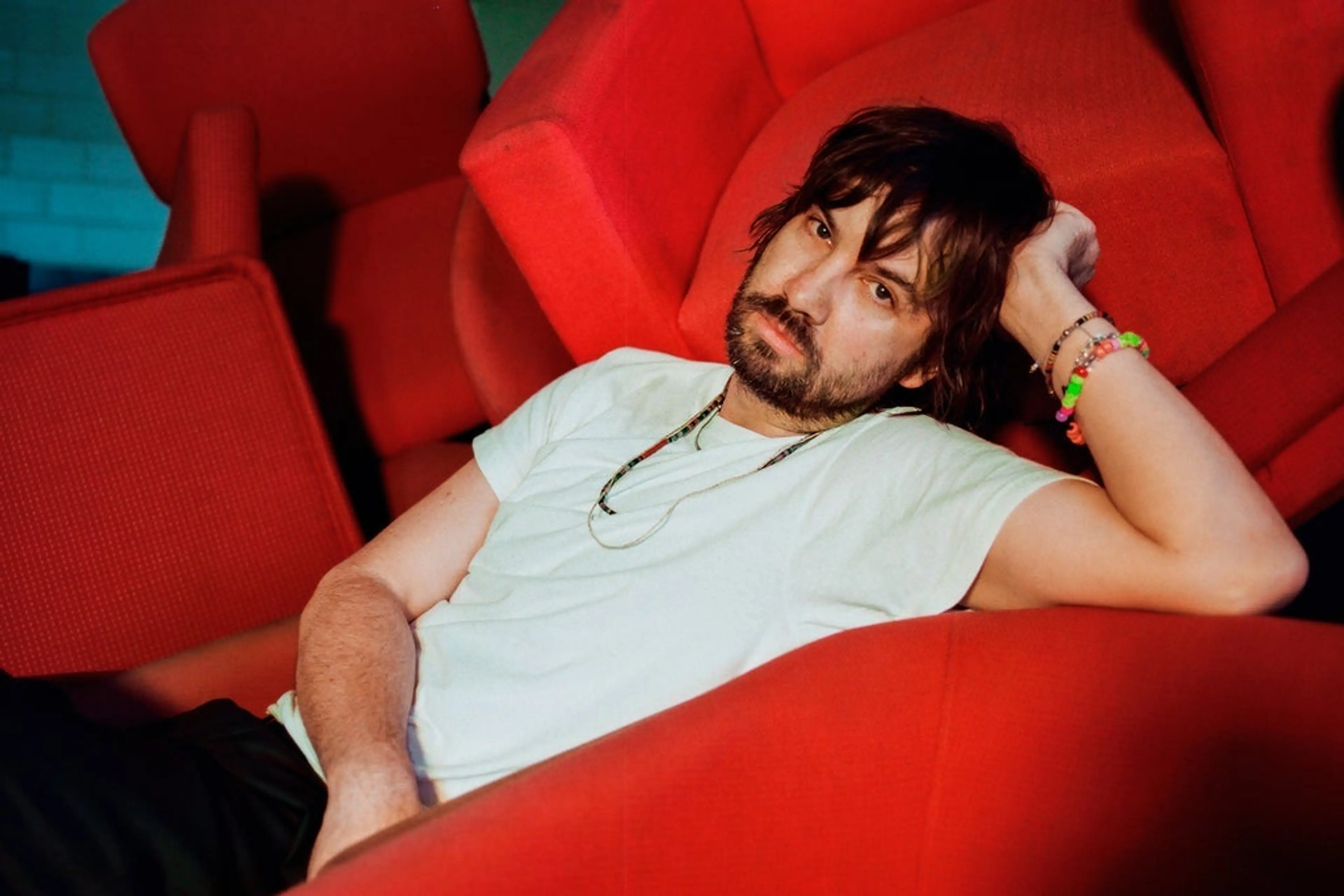 Person with dark hair and beard reclining on red chairs, wearing a white t-shirt and colorful bracelets.