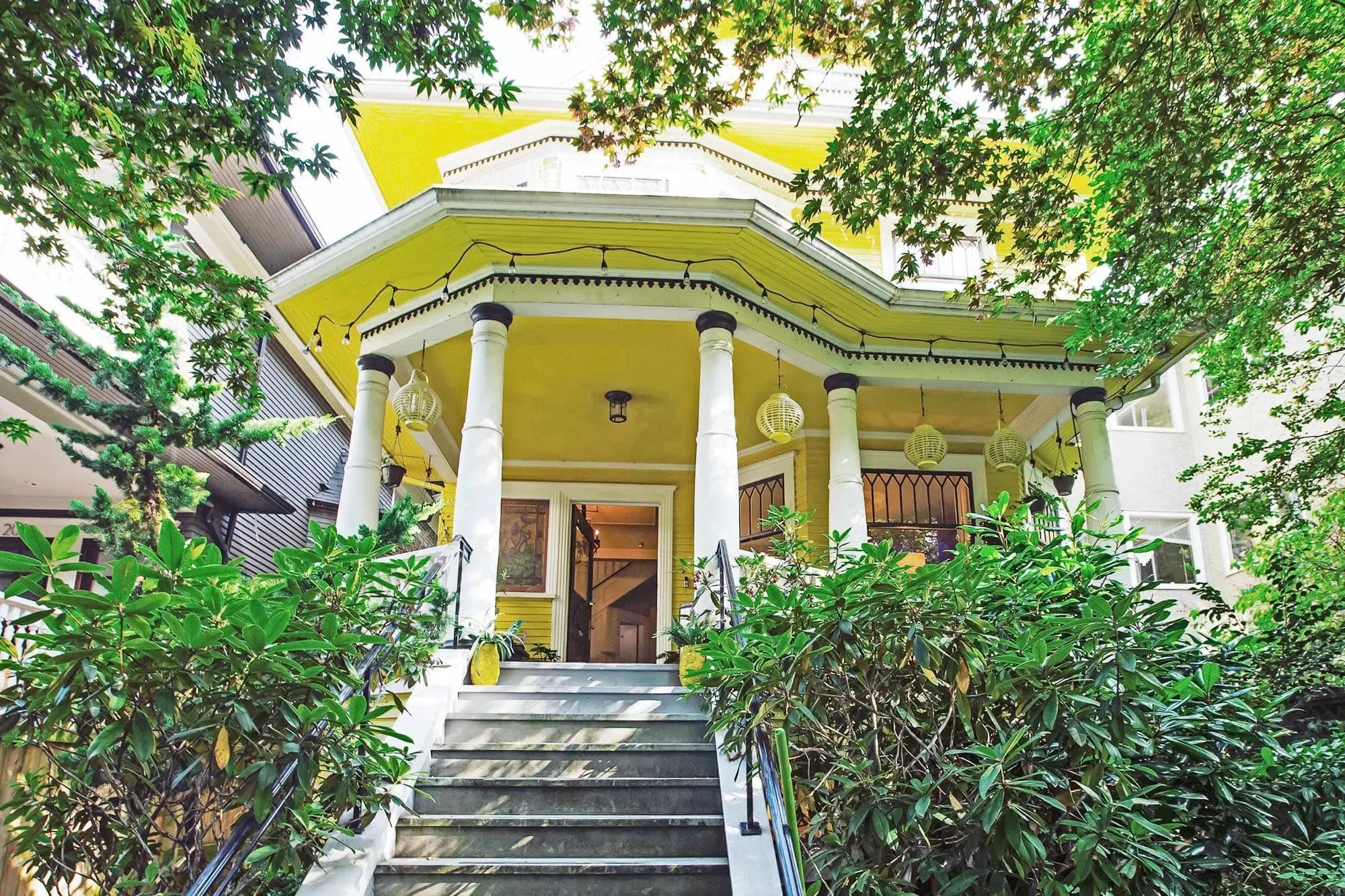 Green plants frame a stone staircase leading up to a bright yellow Victorian-style house