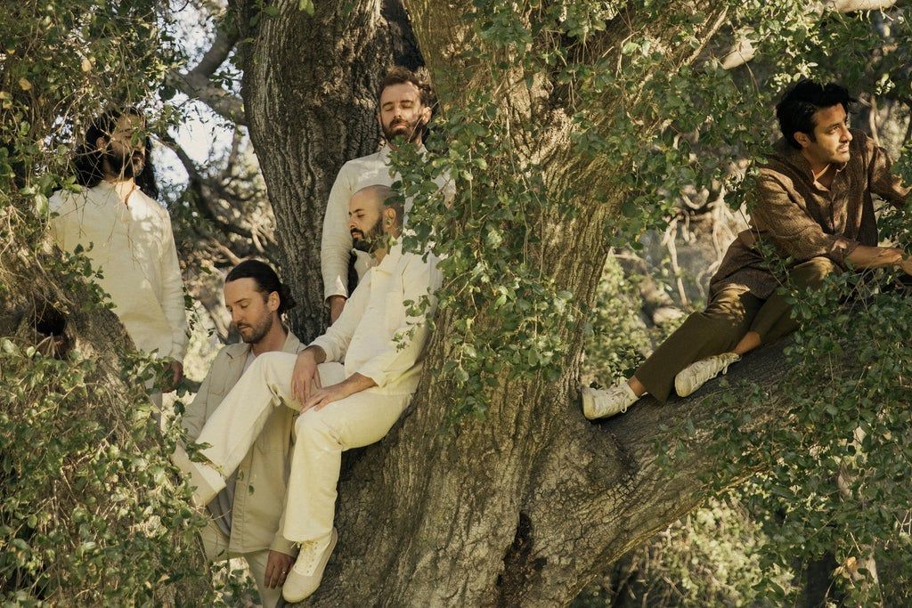 Five people sitting and standing on the branches of a large tree surrounded by green leaves, wearing light and earth-toned clothing.