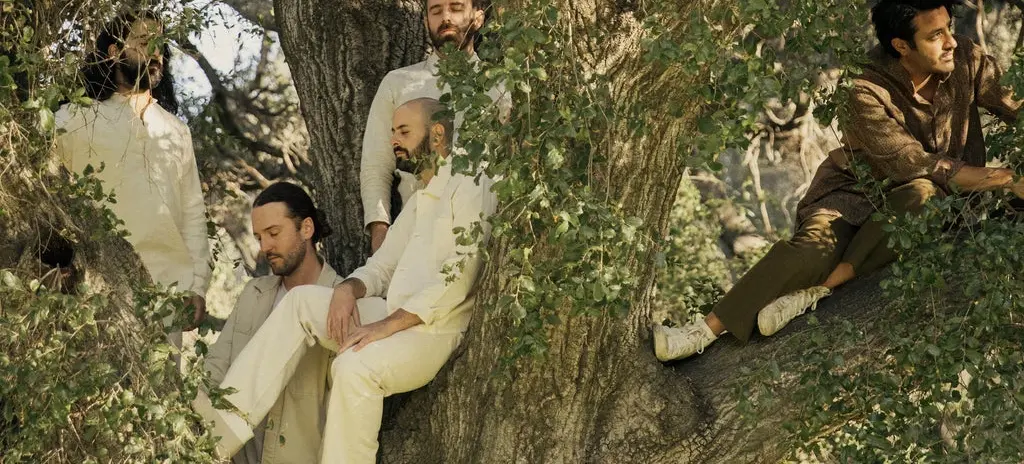 Five people sitting and standing on the branches of a large tree surrounded by green leaves, wearing light and earth-toned clothing.