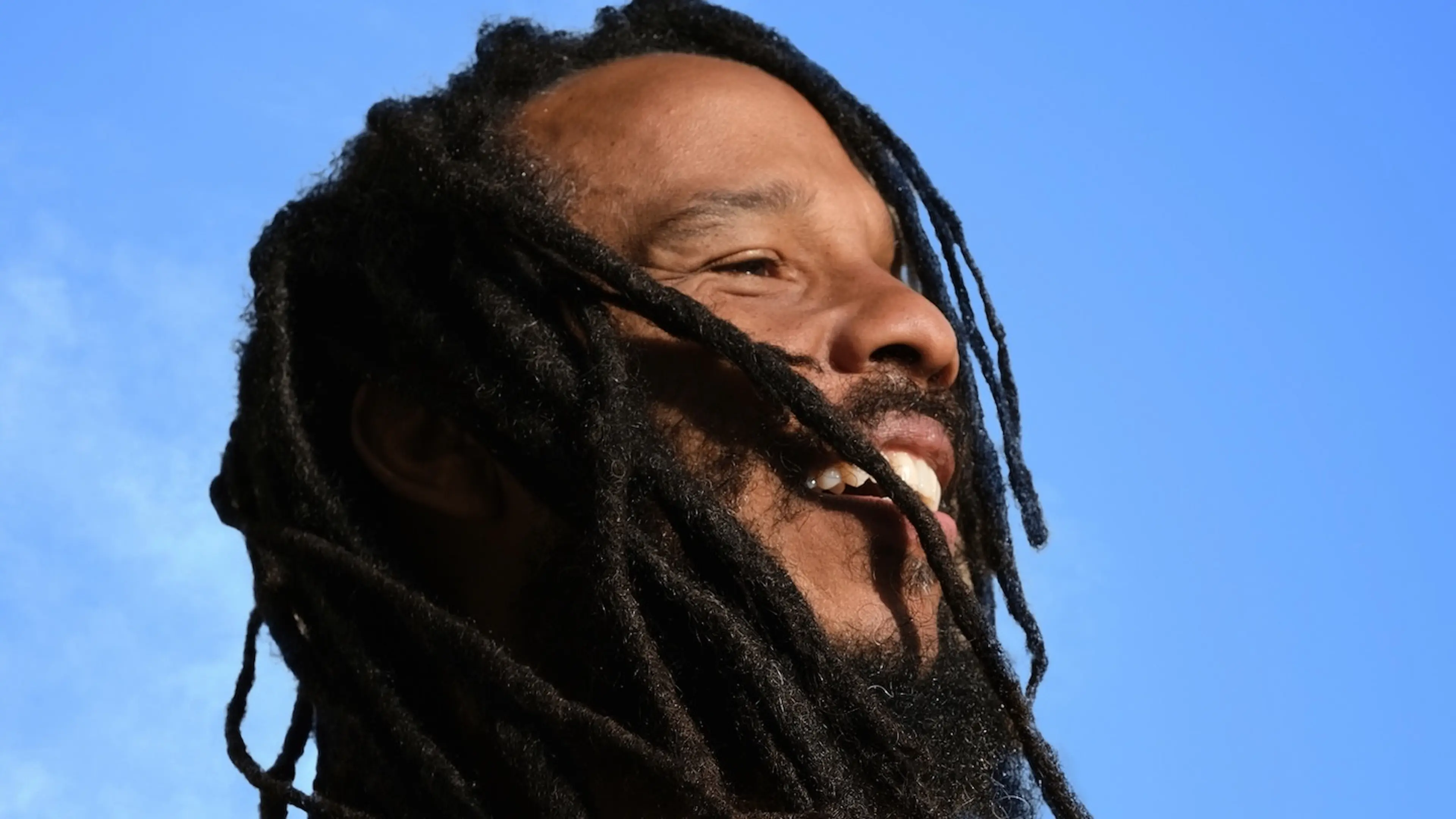 Close-up of a person with long dreadlocks and a beard smiling against a clear blue sky.